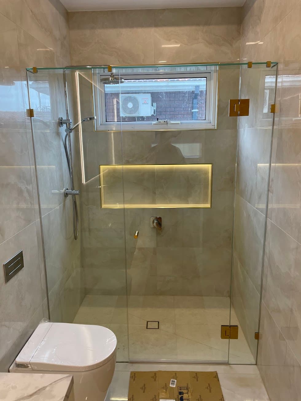 Frameless shower enclosure in a three-panel configuration with brushed gold square hinges and clips mounted directly onto clear glass, installed in a beige marble-tiled bathroom with an illuminated niche.