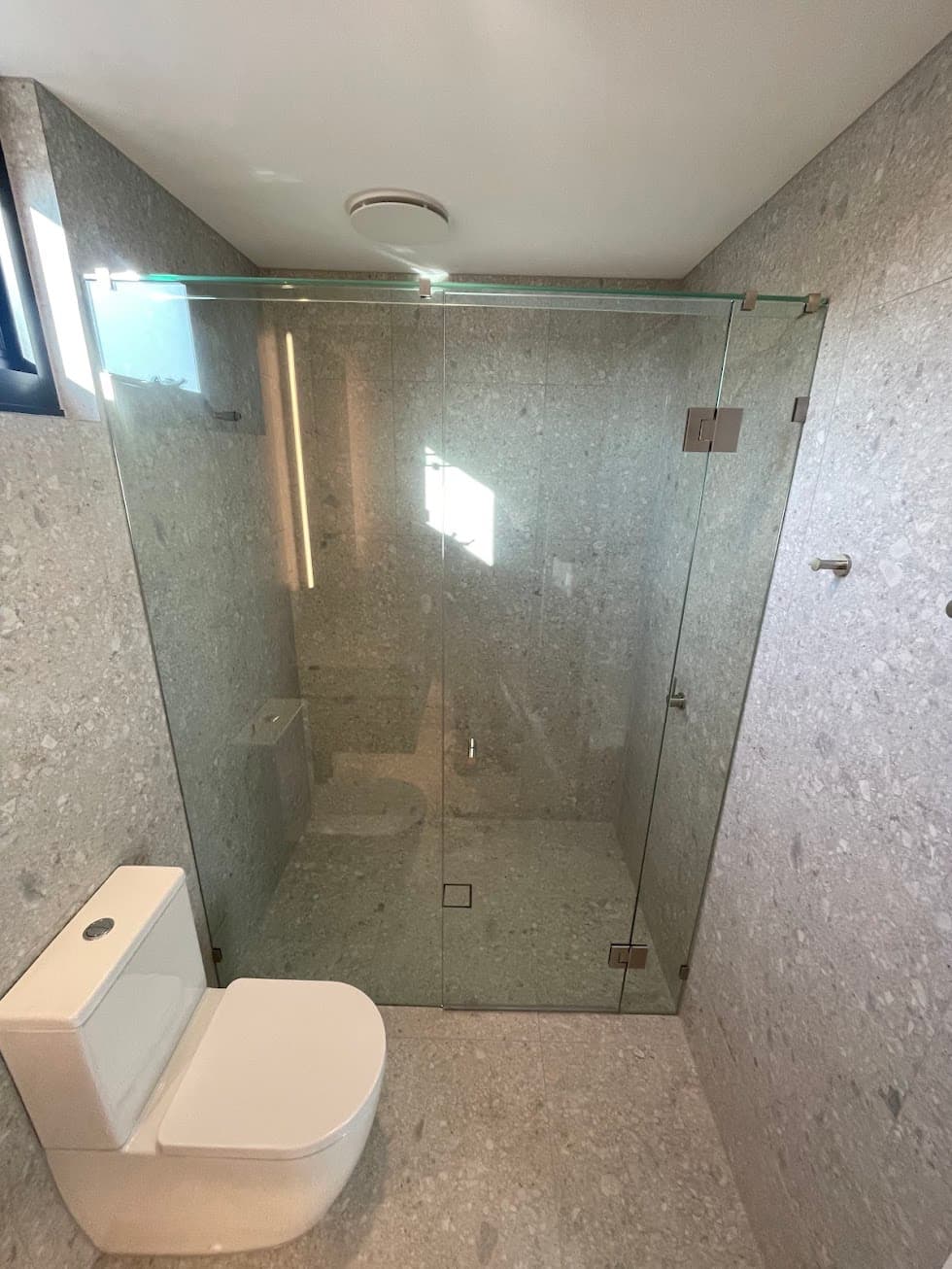 Frameless shower enclosure with a three-panel configuration featuring brushed nickel hinges and hardware, installed in a modern grey terrazzo-tiled bathroom.