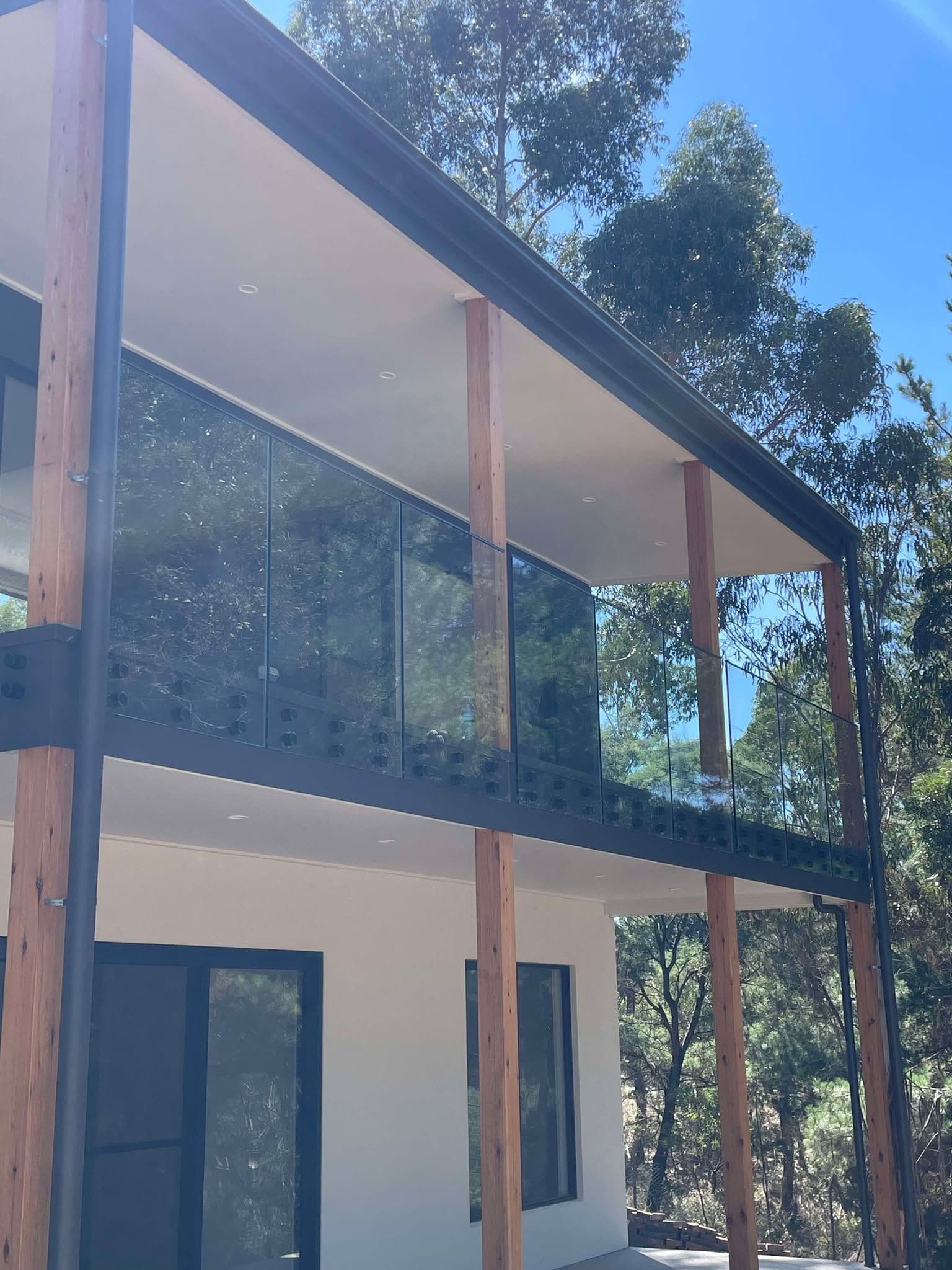 Clear glass balustrade panels fitted to an elevated first-floor balcony with a matte black bottom channel rail, set against a modern timber-post home surrounded by eucalyptus trees.