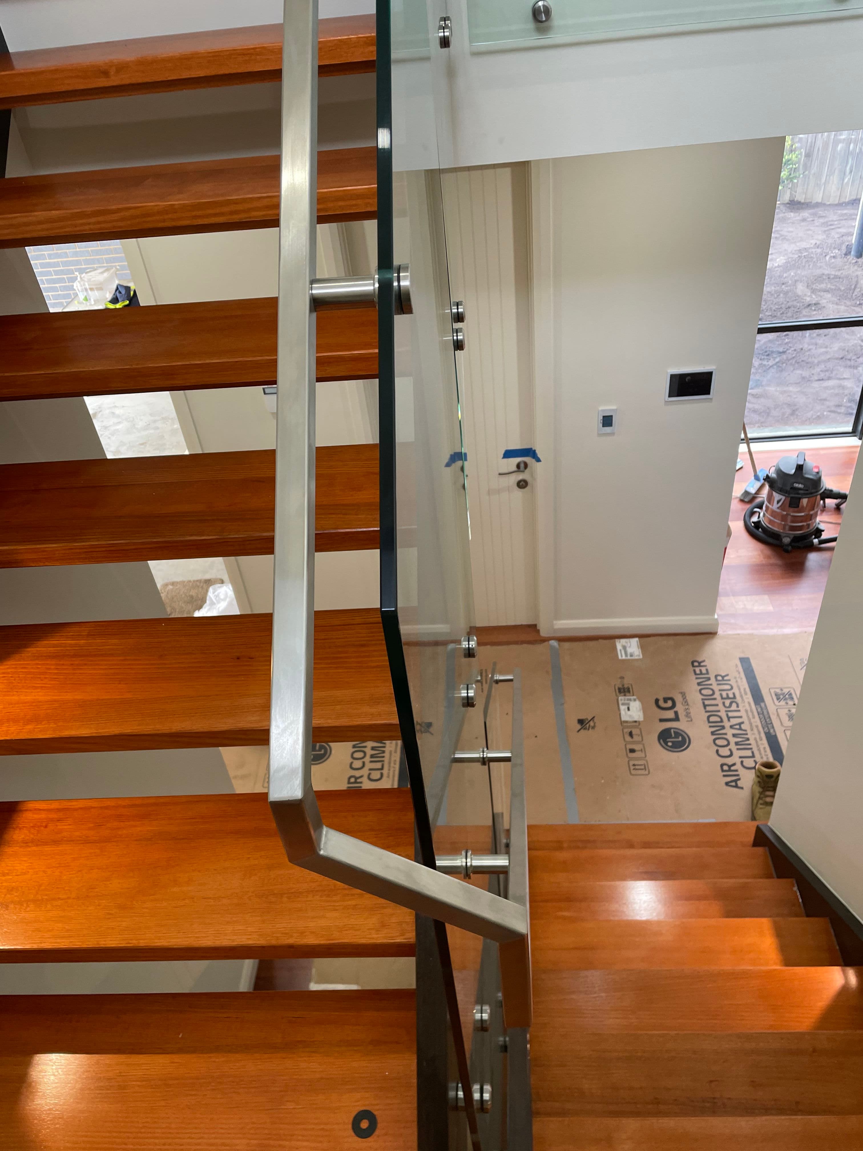 Clear glass staircase balustrade with brushed nickel standoff clamps and handrail channel alongside timber open-tread stairs in a residential interior under construction.