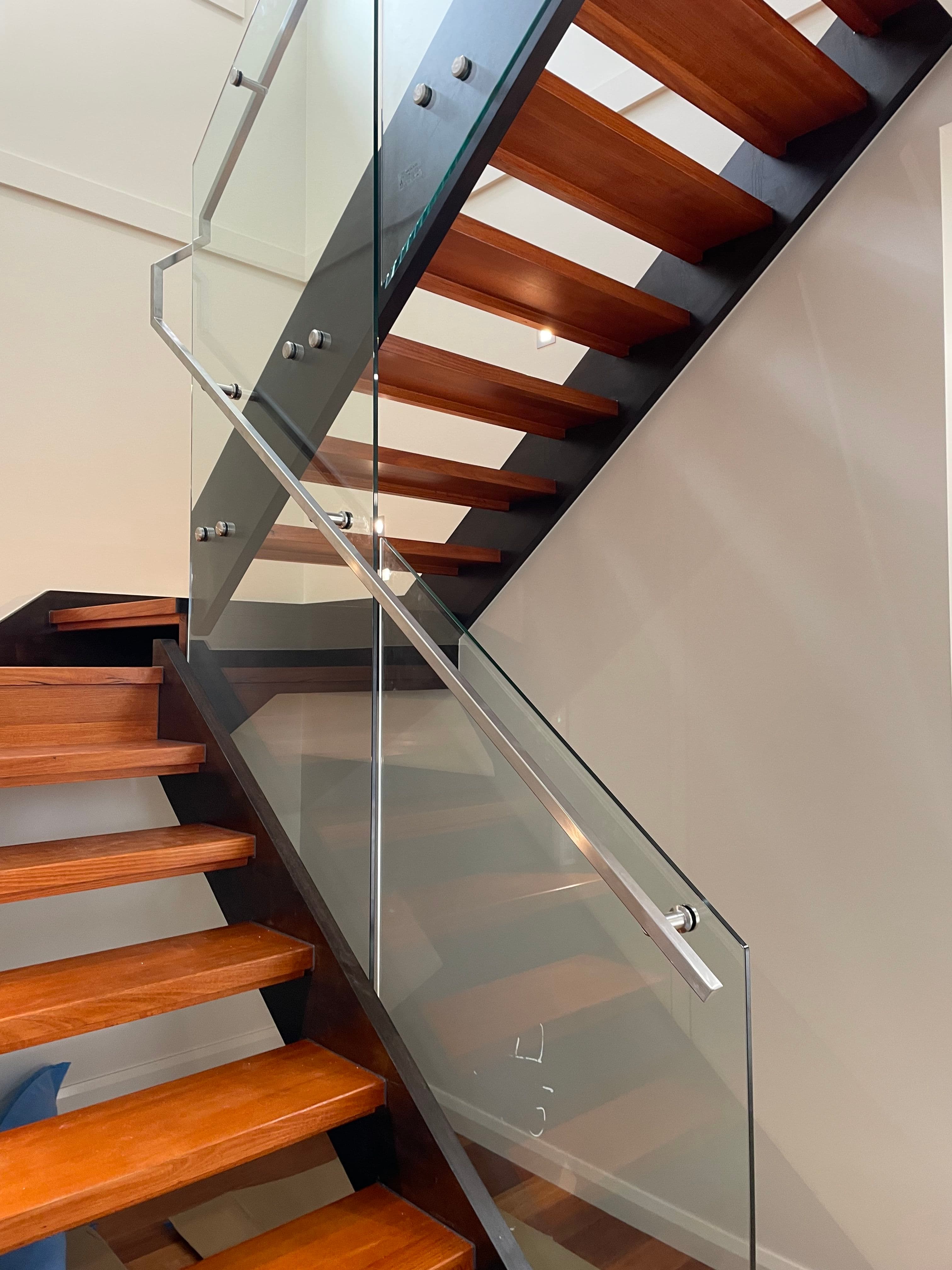 Clear glass stair balustrade with brushed nickel standoff fixings mounted to a dark steel stringer on open-tread timber stairs in a modern interior.
