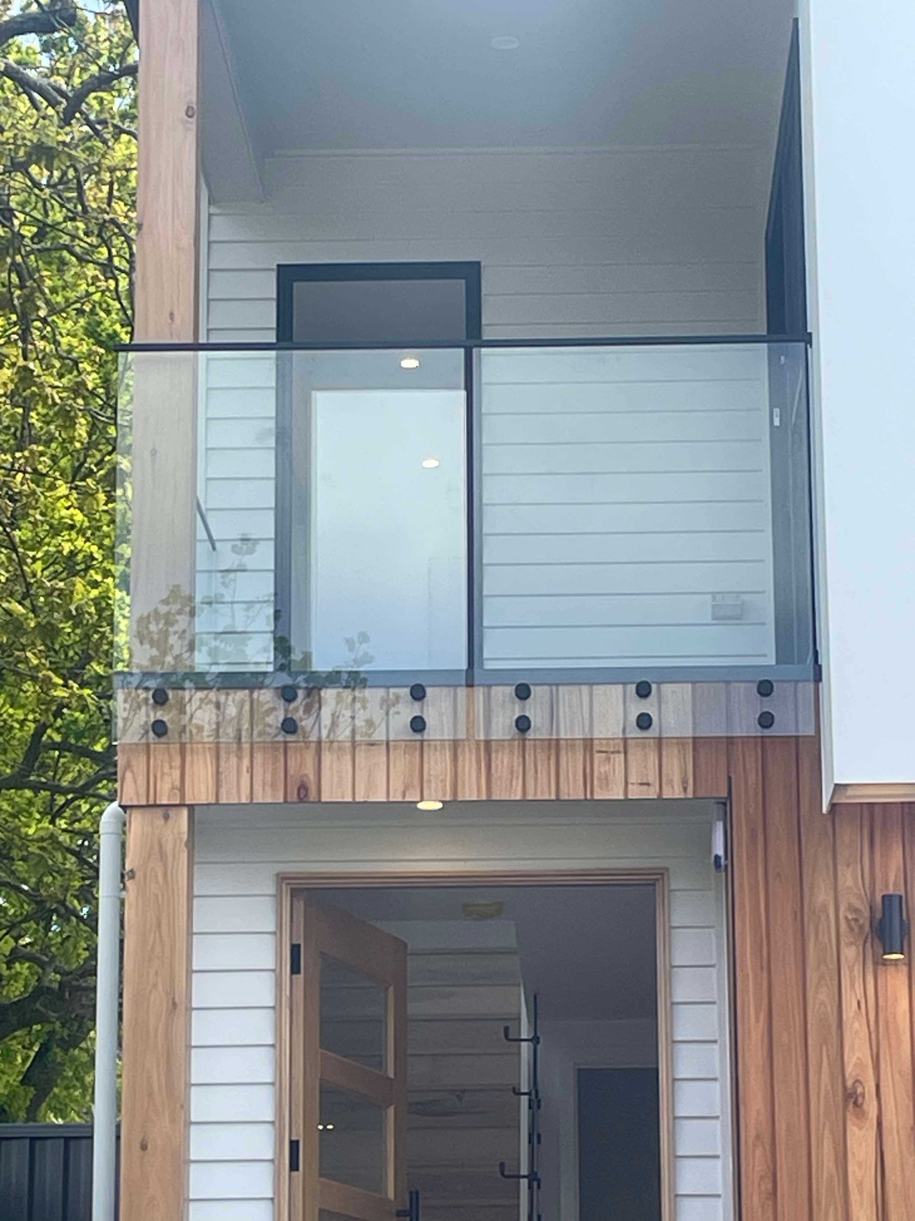 Clear glass balustrade panels with matte black top rail and button spigots fixed to a timber-clad first-floor balcony on a contemporary two-storey home exterior.
