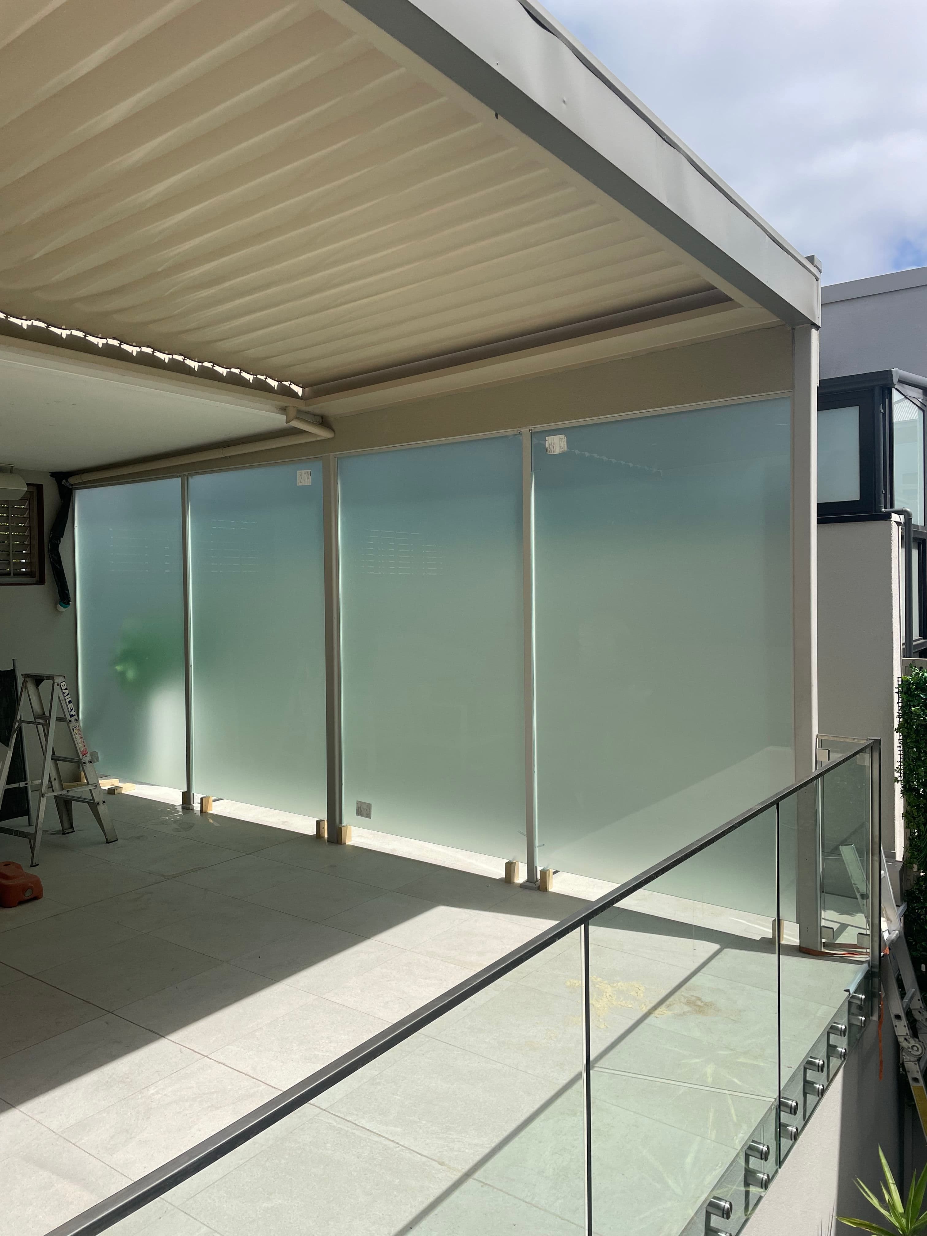 Frameless glass balustrade with chrome spigots on an elevated outdoor terrace, with acid etched glass privacy panels fitted beneath a louvred pergola in the background.