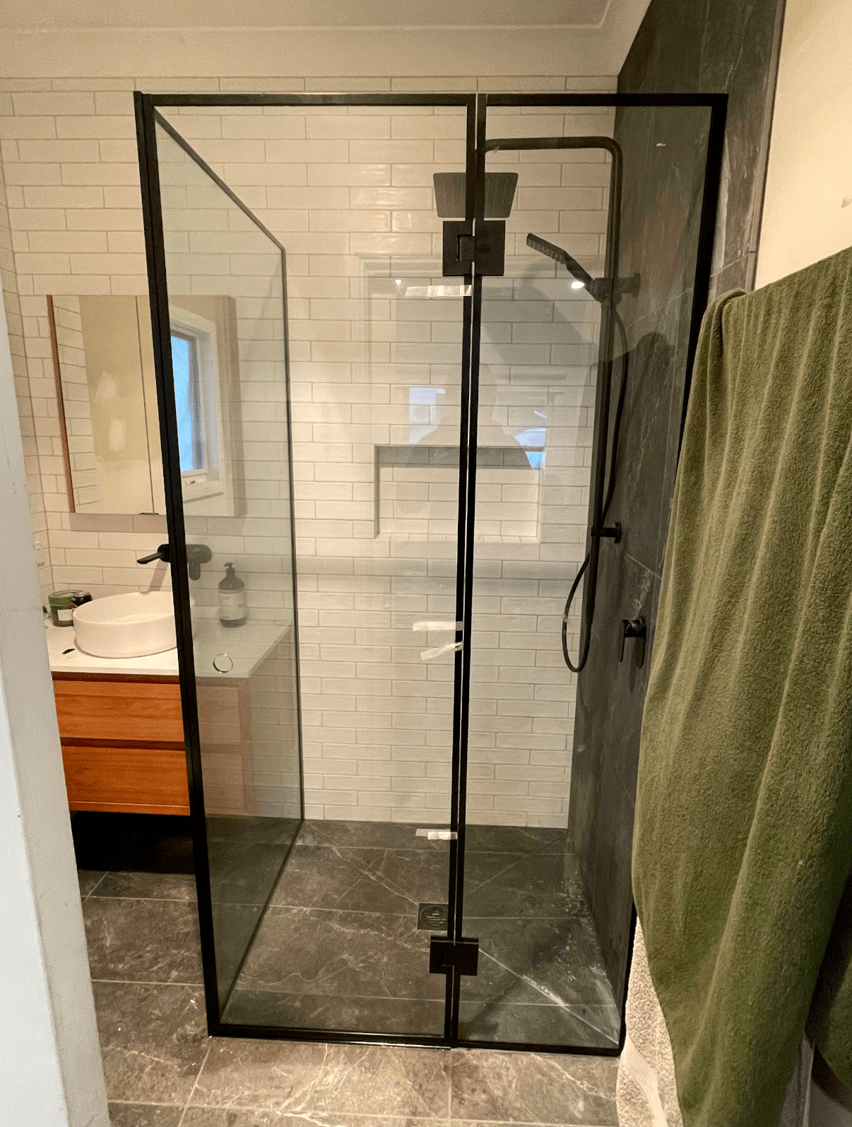 Semi-frameless shower enclosure with full matte black aluminium frame rails on a corner configuration with hinged door, set in a tiled bathroom with subway tiles and marble-look floor tiles.
