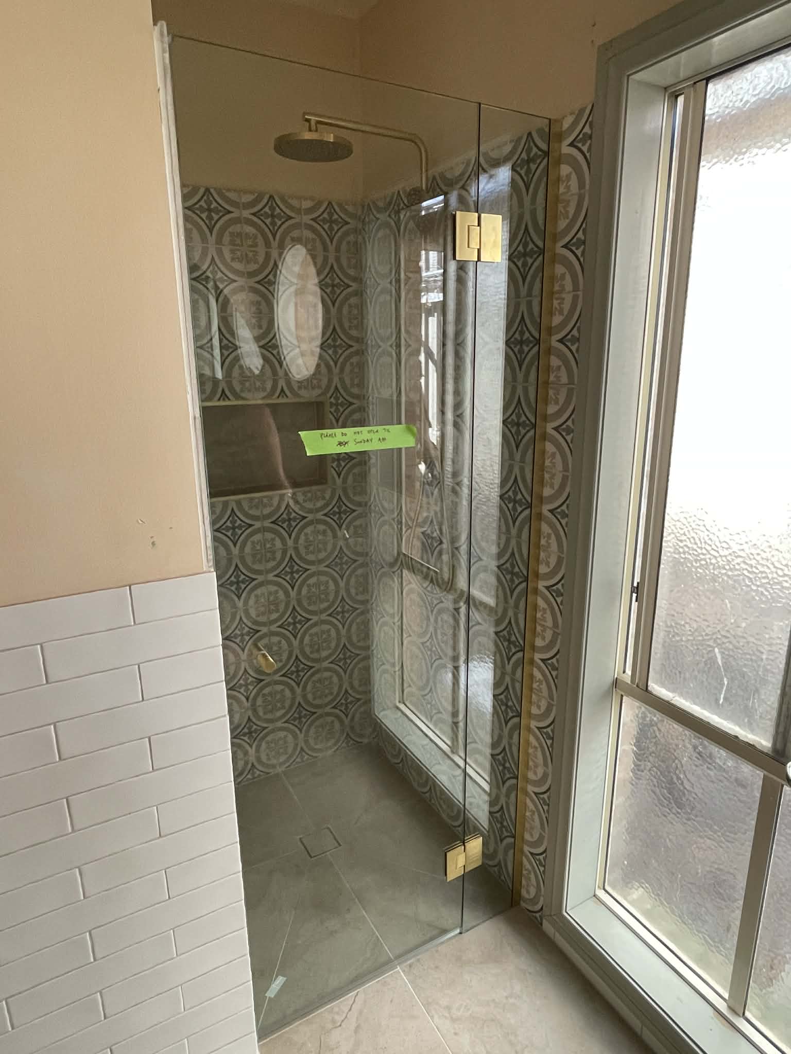 Frameless shower enclosure with brushed gold hinges and pivot hardware on a two-panel configuration in a patterned-tile bathroom with subway tile surround.