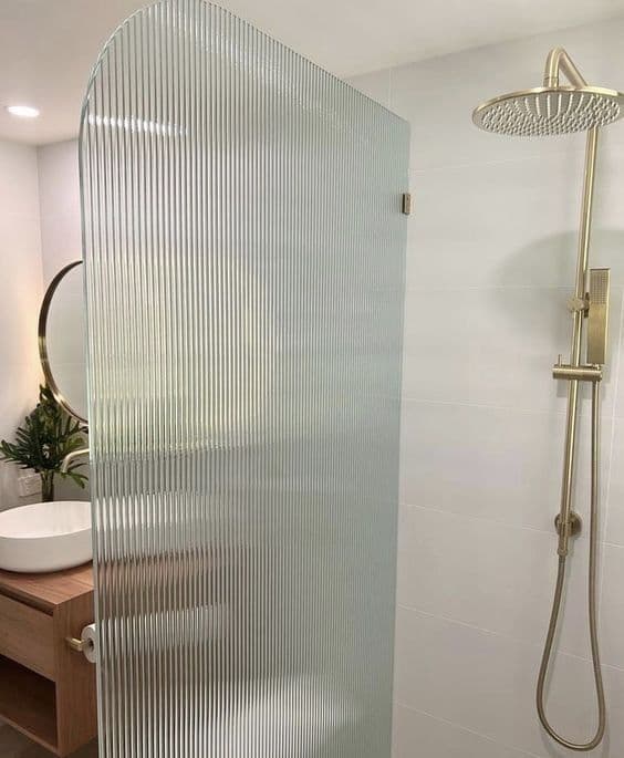Frameless reeded glass shower screen with an arched top profile and brushed gold hardware in a white tiled bathroom with a timber vanity.