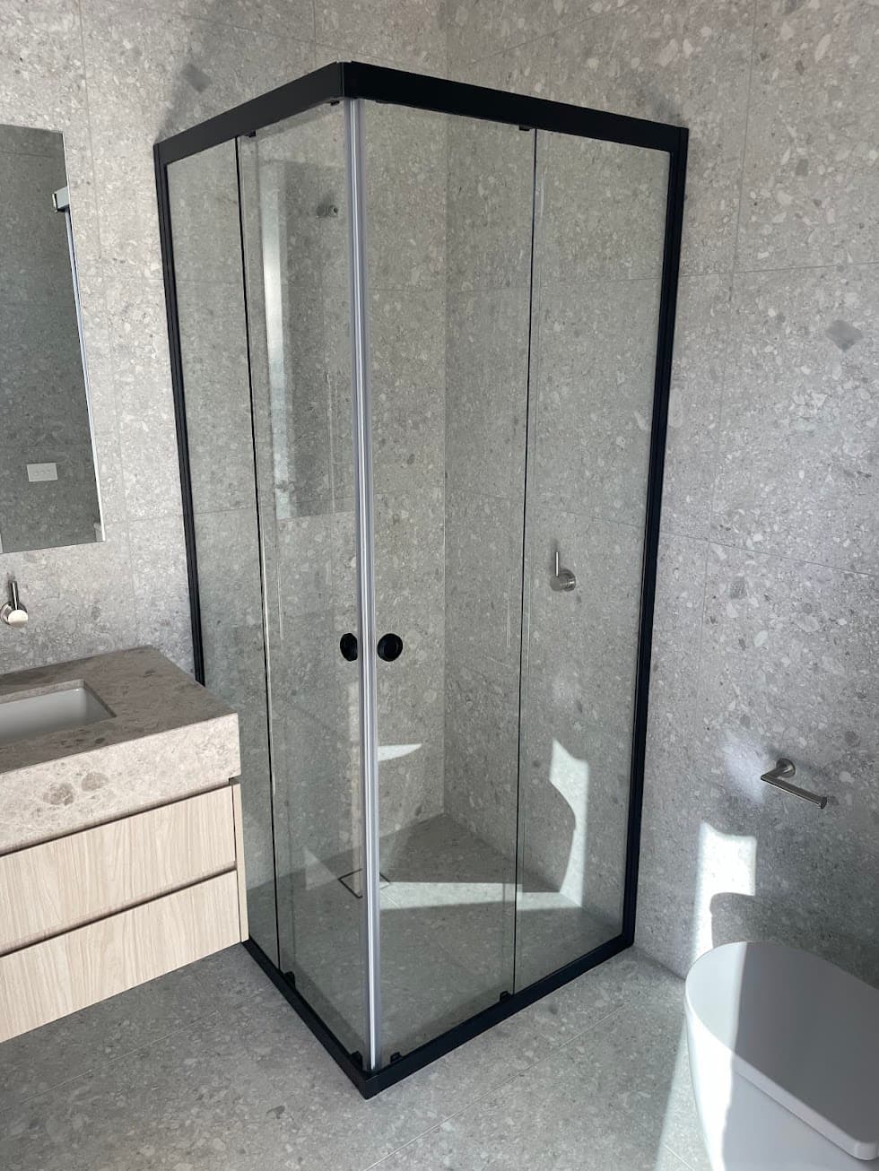 Semi-frameless corner shower enclosure with matte black aluminium frame rails, top channel, and base track featuring sliding doors with round pull handles in a grey stone-tiled bathroom.