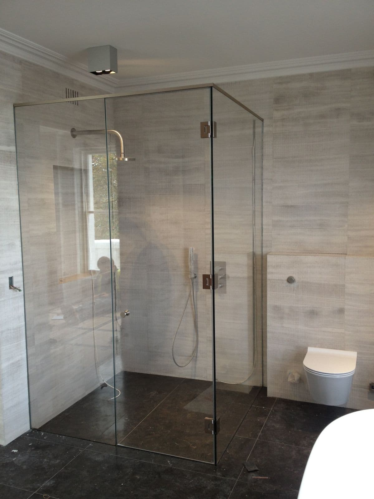 Frameless shower enclosure with a door hung off a return panel, featuring brushed nickel square hinges and pivot hardware, clear glass in a contemporary tiled bathroom with dark stone floor tiles.