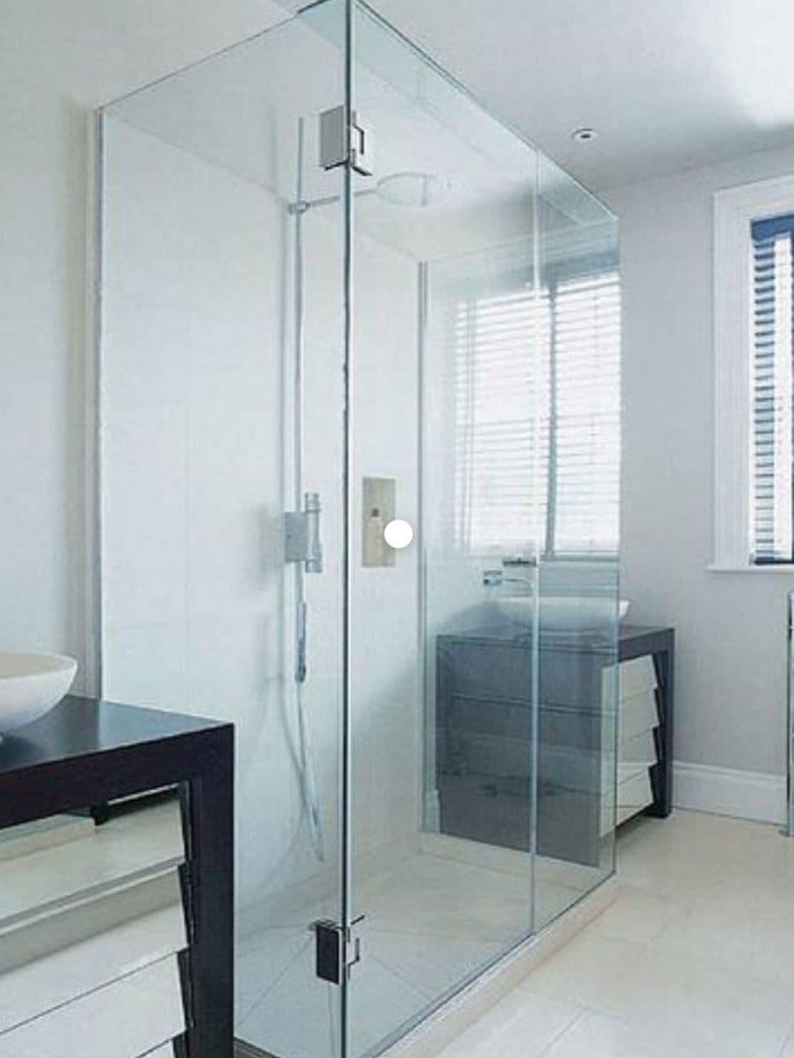 Frameless shower enclosure in a corner configuration with a door hung off a return panel, featuring chrome hinges and hardware in a bright white bathroom with large format tiles.
