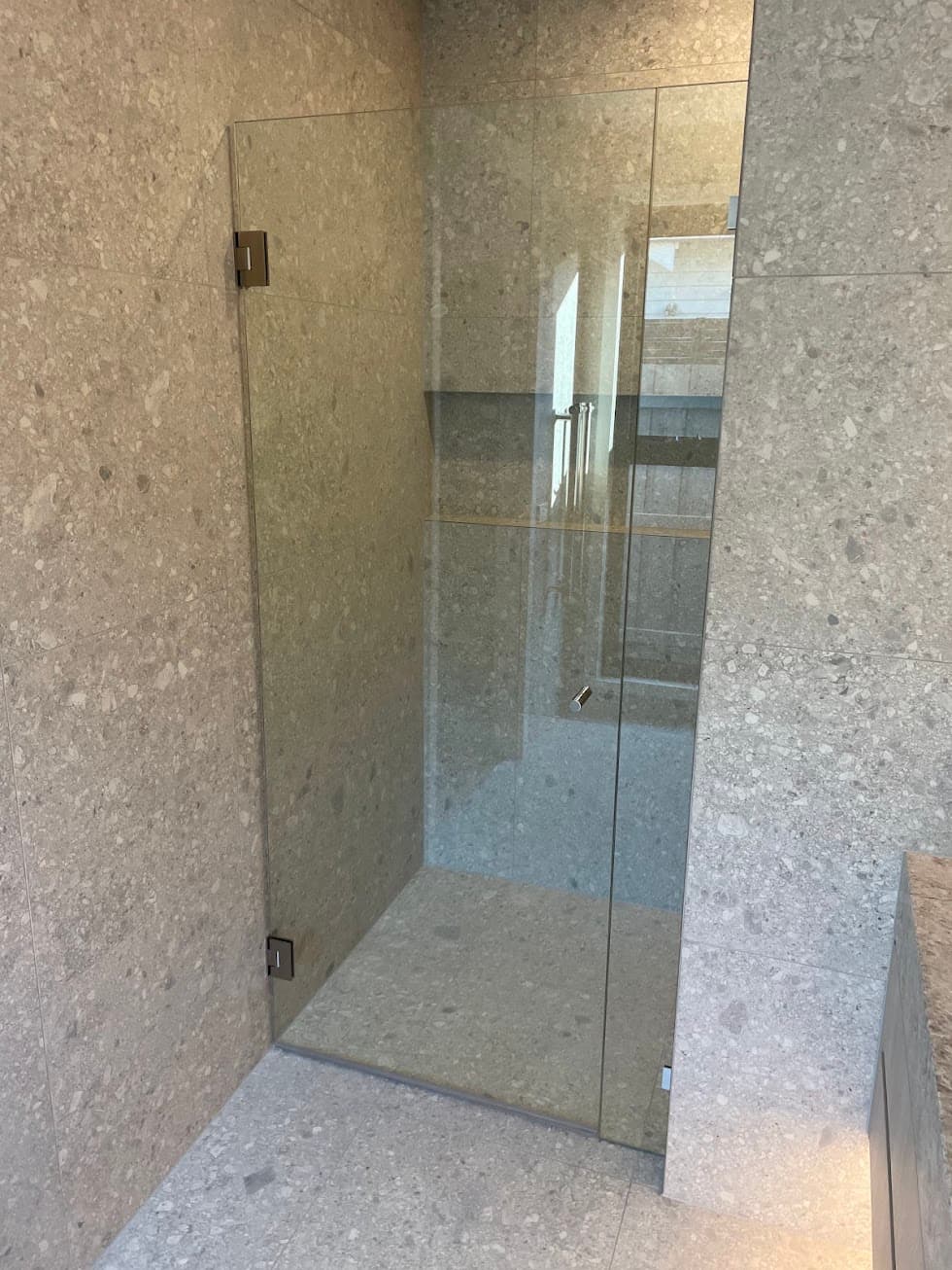 Frameless shower door hinged directly off the wall with brushed nickel wall-mounted hinges, clear glass panel in a floor-to-ceiling stone-tiled ensuite with no aluminium frame or channel visible.