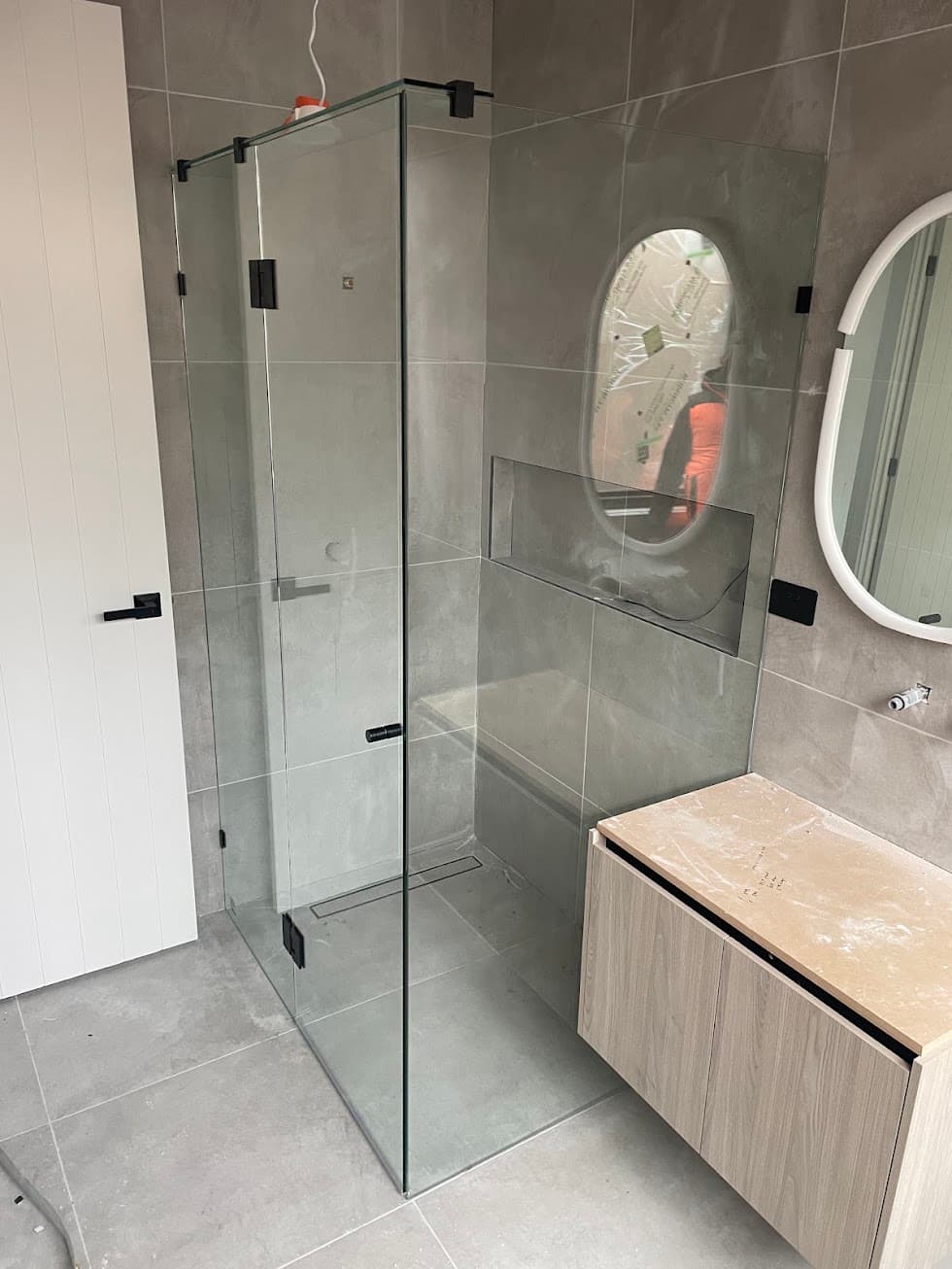 Frameless shower enclosure in a front-return configuration with matte black hinges and hardware mounted directly onto clear glass panels, installed in a modern grey-tiled bathroom.