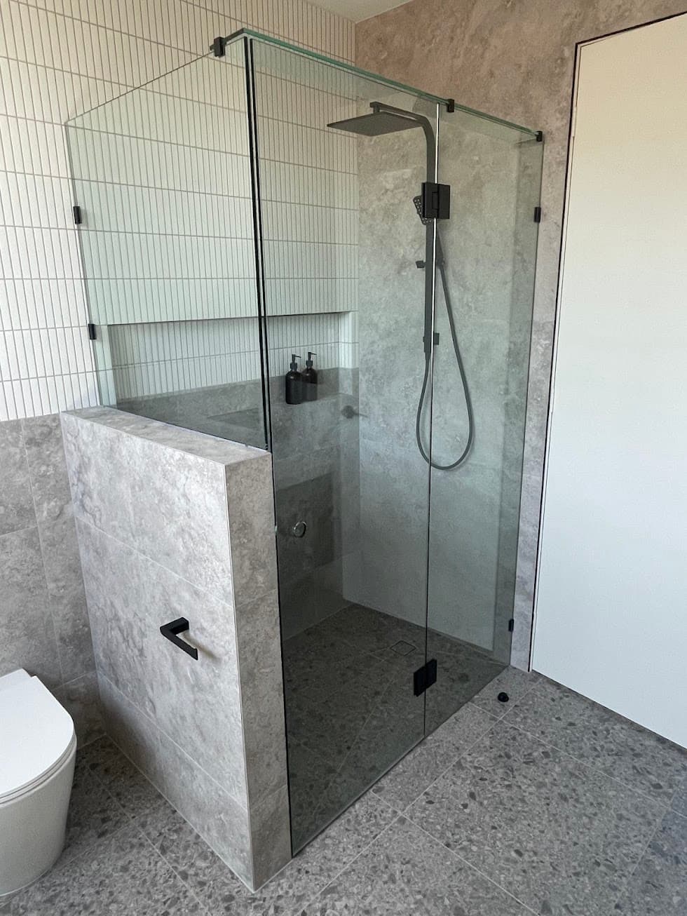 Frameless shower enclosure with a front-and-return configuration mounted off a tiled nib wall, featuring matte black hinges and hardware in a modern grey stone-tiled bathroom.