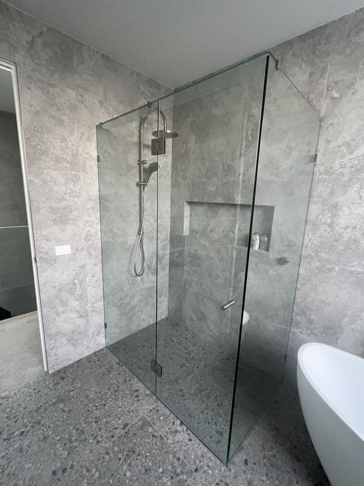Frameless shower enclosure in a front-return configuration with brushed nickel hinges and pivot hardware, clear glass panels mounted directly on the glass edge, in a contemporary bathroom with large-format grey stone-look tiles and a freestanding bath.