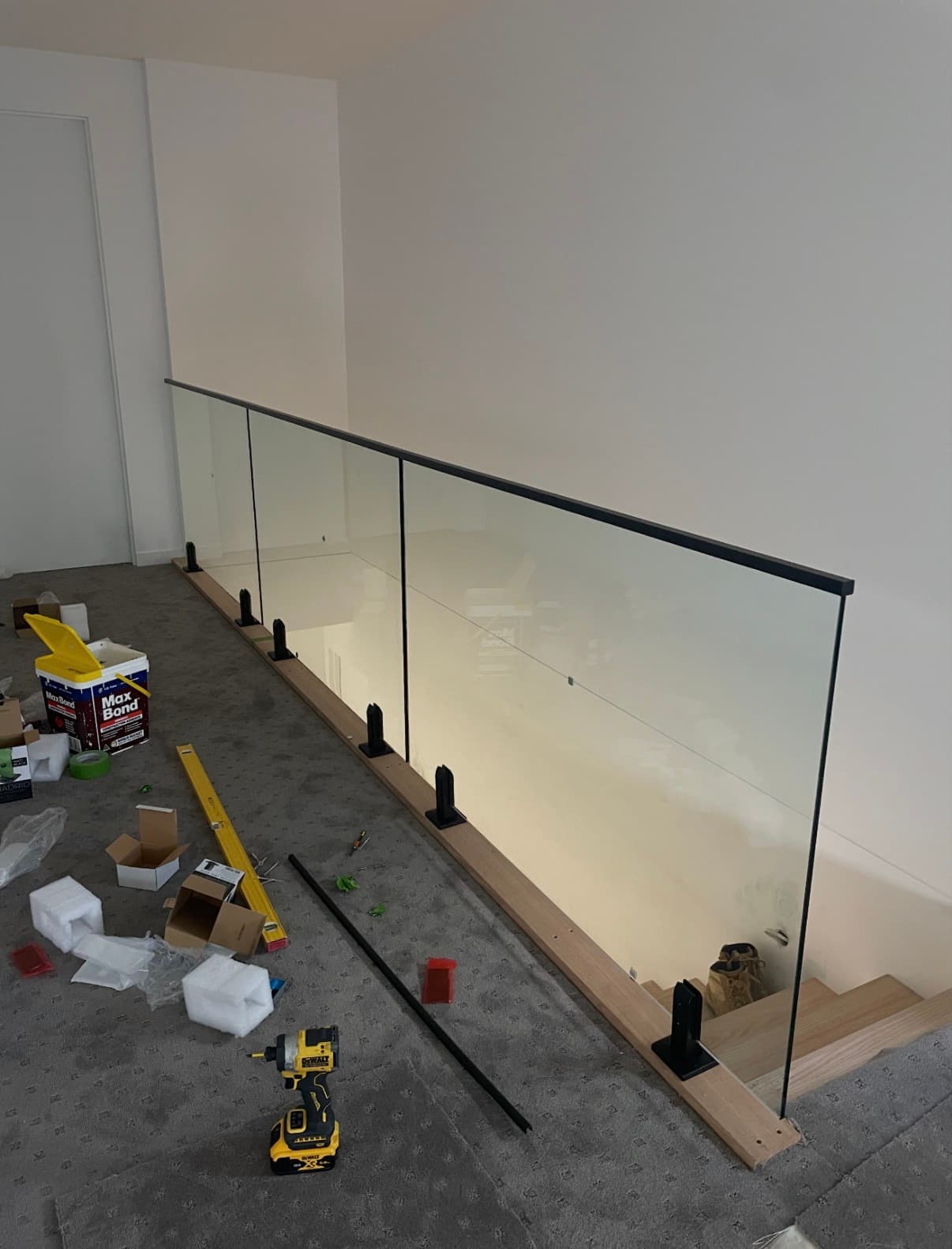 Clear glass balustrade with matte black spigots and top handrail installed along a timber-edged staircase landing in a residential interior, photographed mid-installation with tools and materials visible.