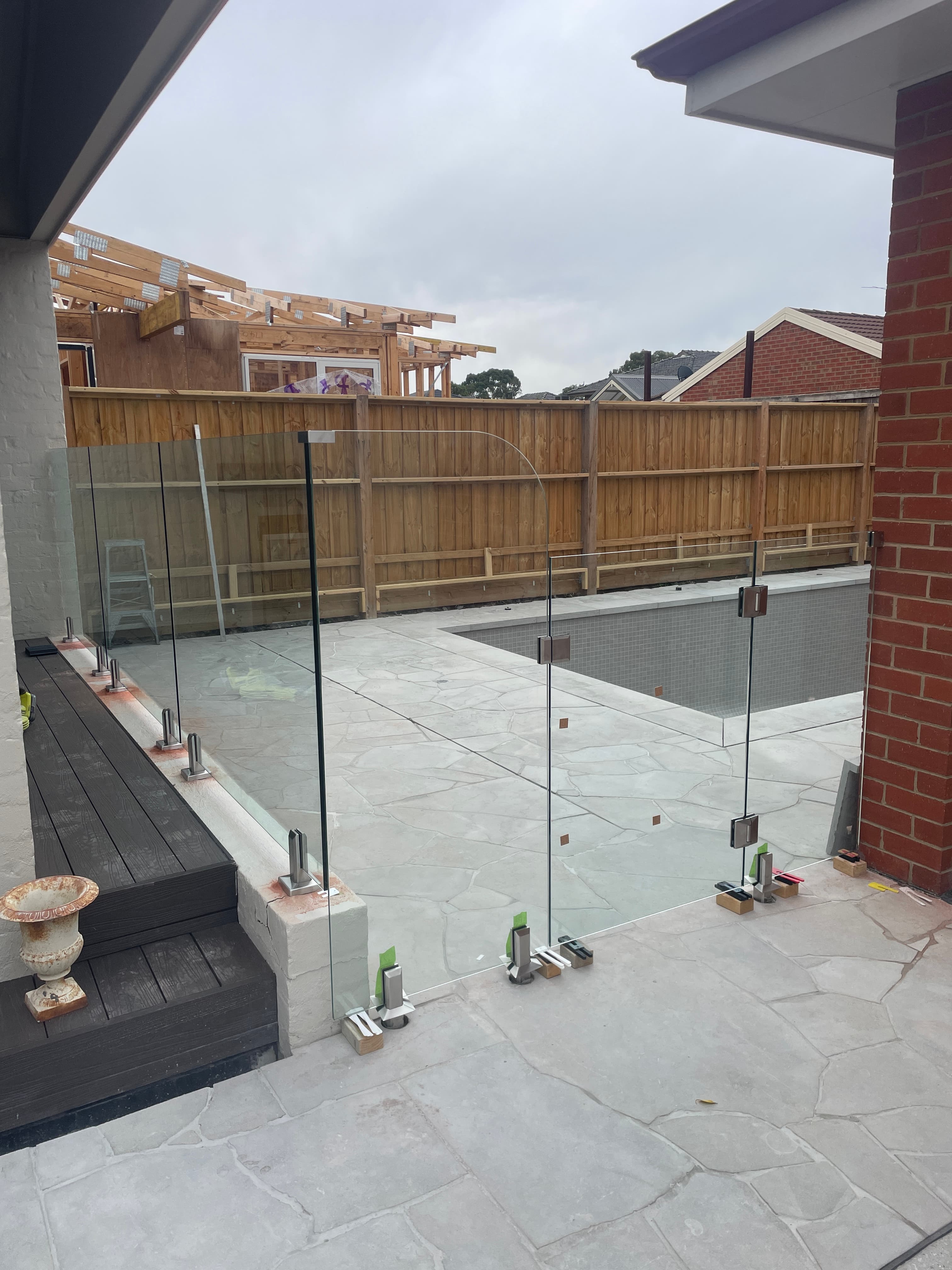 Frameless pool fence with brushed chrome spigots core-drilled into stone coping surrounding an under-construction backyard swimming pool, viewed from a paved alfresco area.