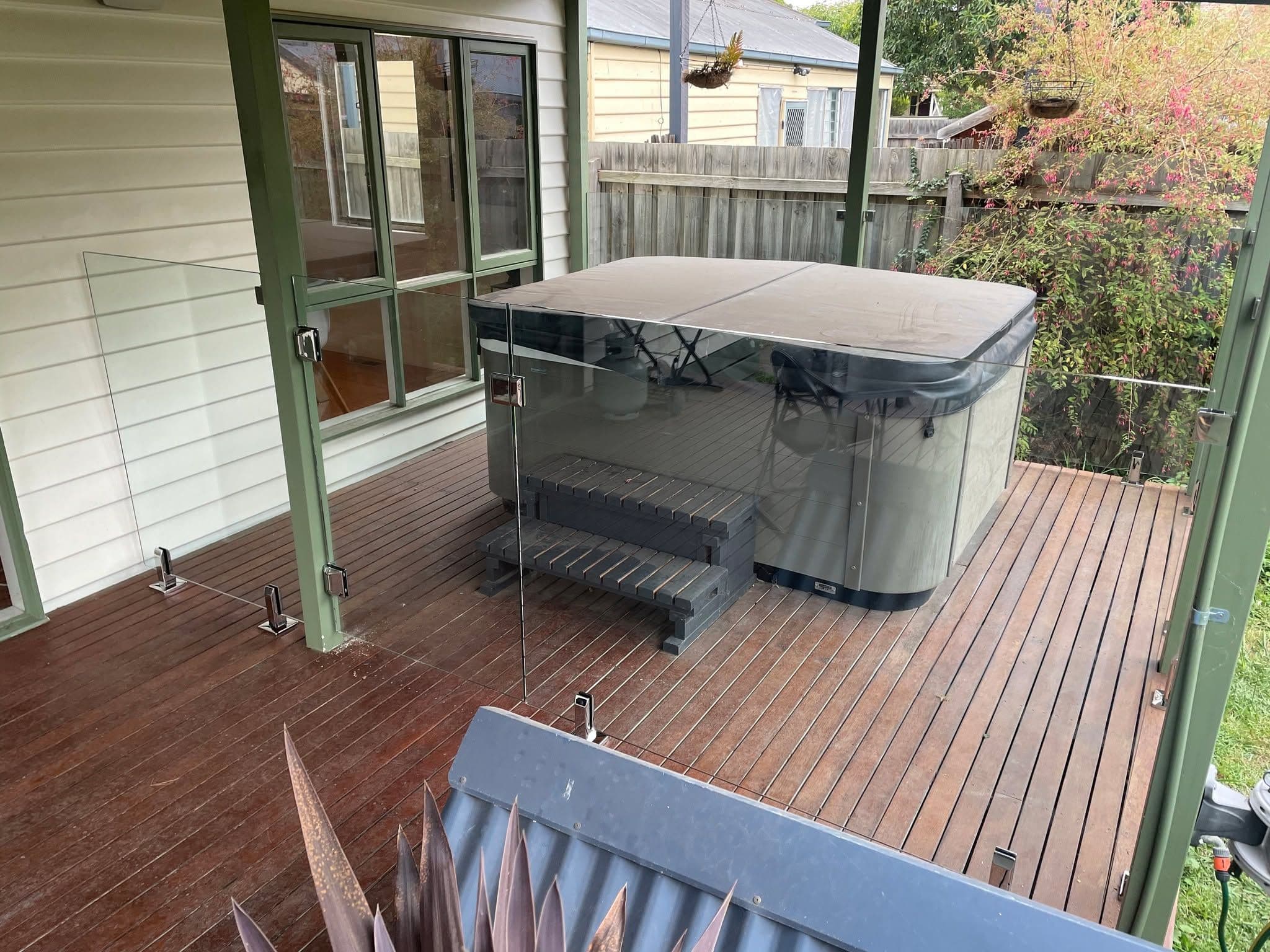 Frameless glass balustrade with chrome spigots installed on a timber deck surrounding a covered spa/hot tub beside a weatherboard house exterior.
