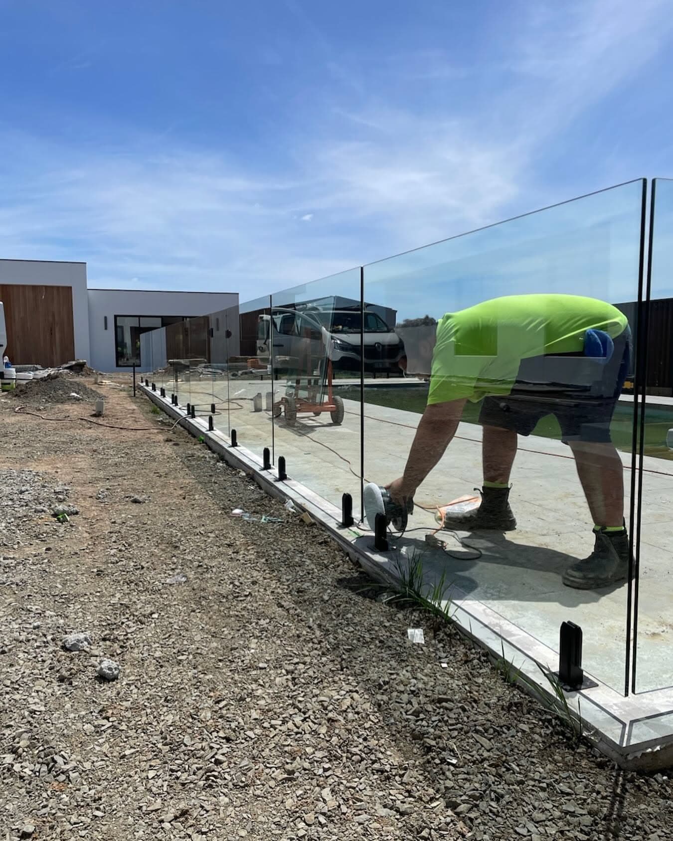 Clear glass pool fence panels being installed with matte black spigots core-drilled into concrete coping alongside a residential pool under construction.