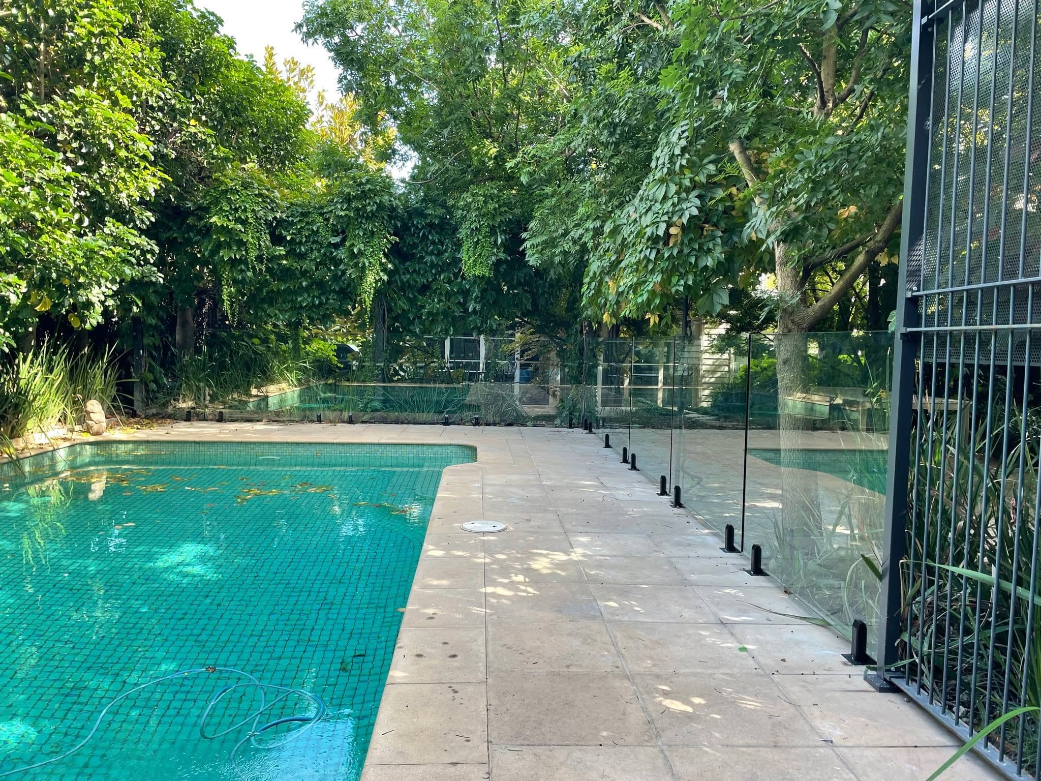 Frameless glass pool fence with matte black spigots core-drilled into concrete coping surrounding a backyard swimming pool, set in a lush garden.