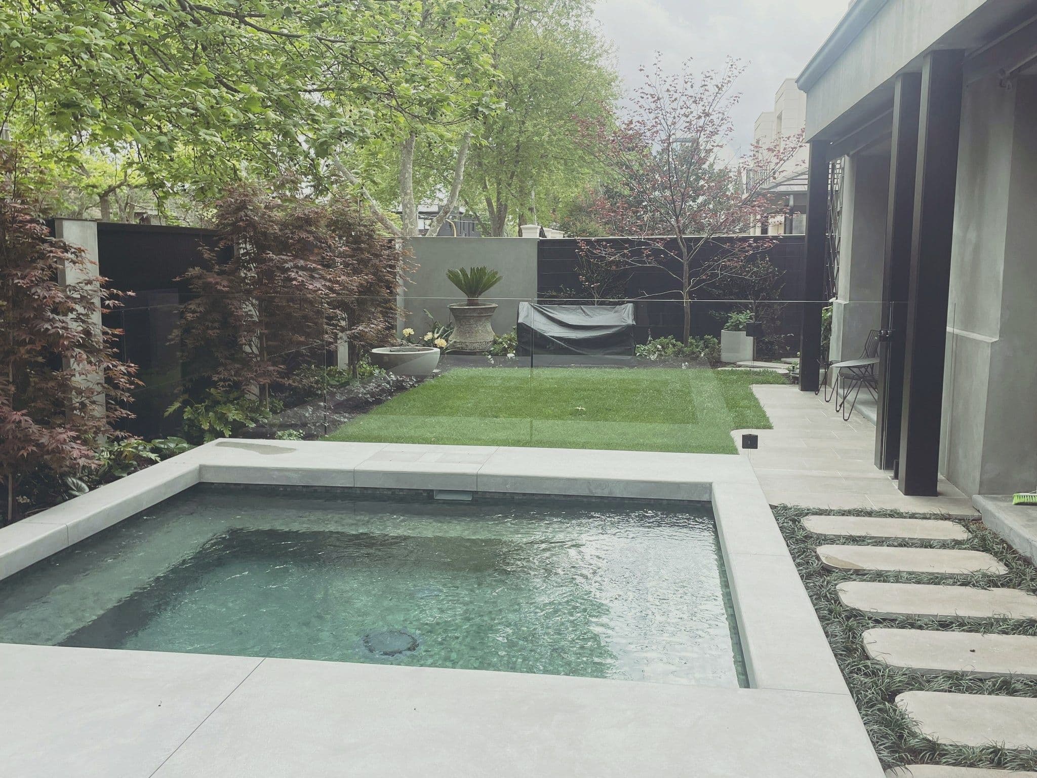Frameless glass pool fence with spigots installed around a compact plunge pool with light stone coping in a landscaped backyard garden setting.