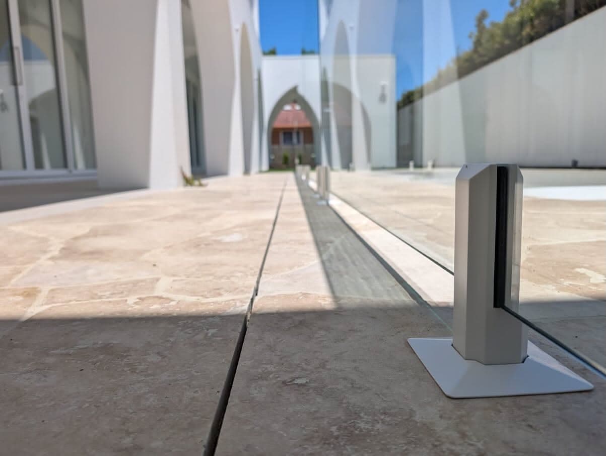 Frameless glass balustrade with brushed chrome square spigots surface-mounted on travertine stone paving at an outdoor terrace or courtyard of a modern white residential property.