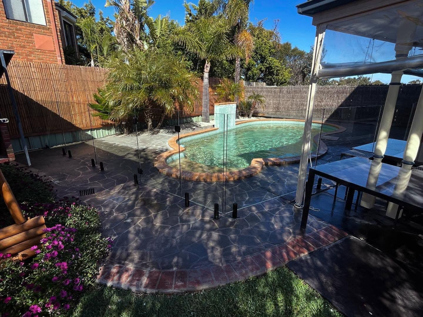 Frameless glass pool fence with matte black spigots core-drilled into a patterned concrete and brick coping surrounding a freeform backyard swimming pool with palm trees and garden landscaping.