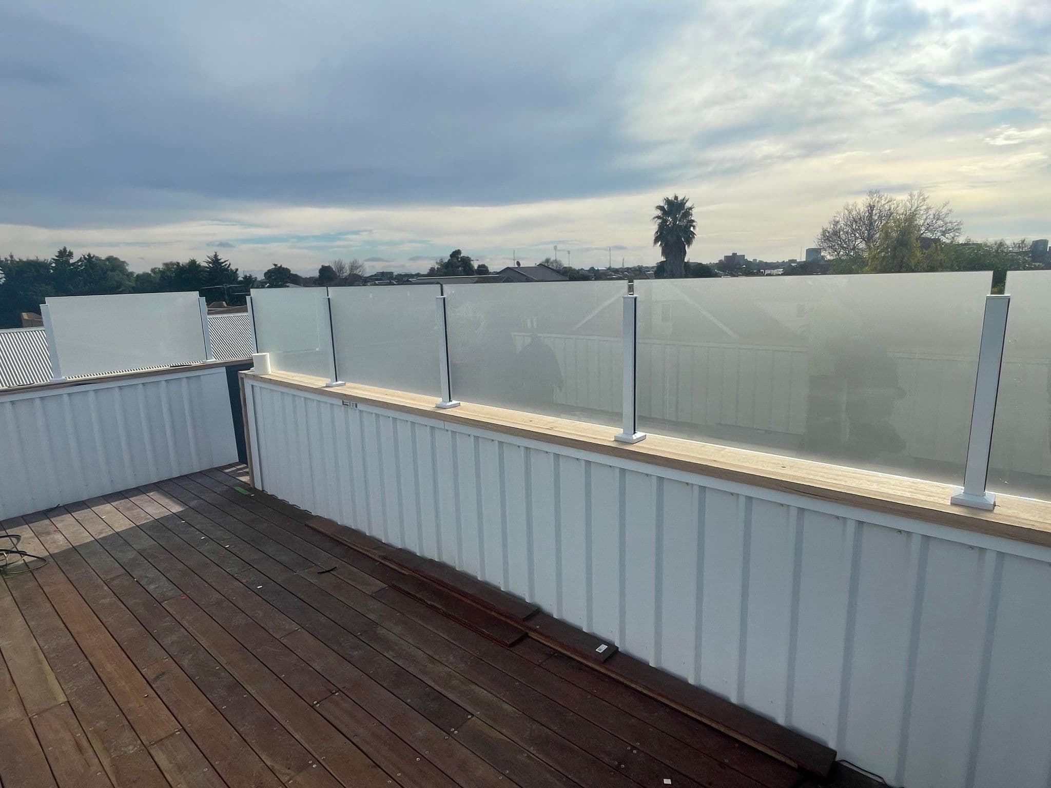 Acid etched glass balustrade panels with white square spigots installed on a timber deck rooftop terrace overlooking a suburban skyline.