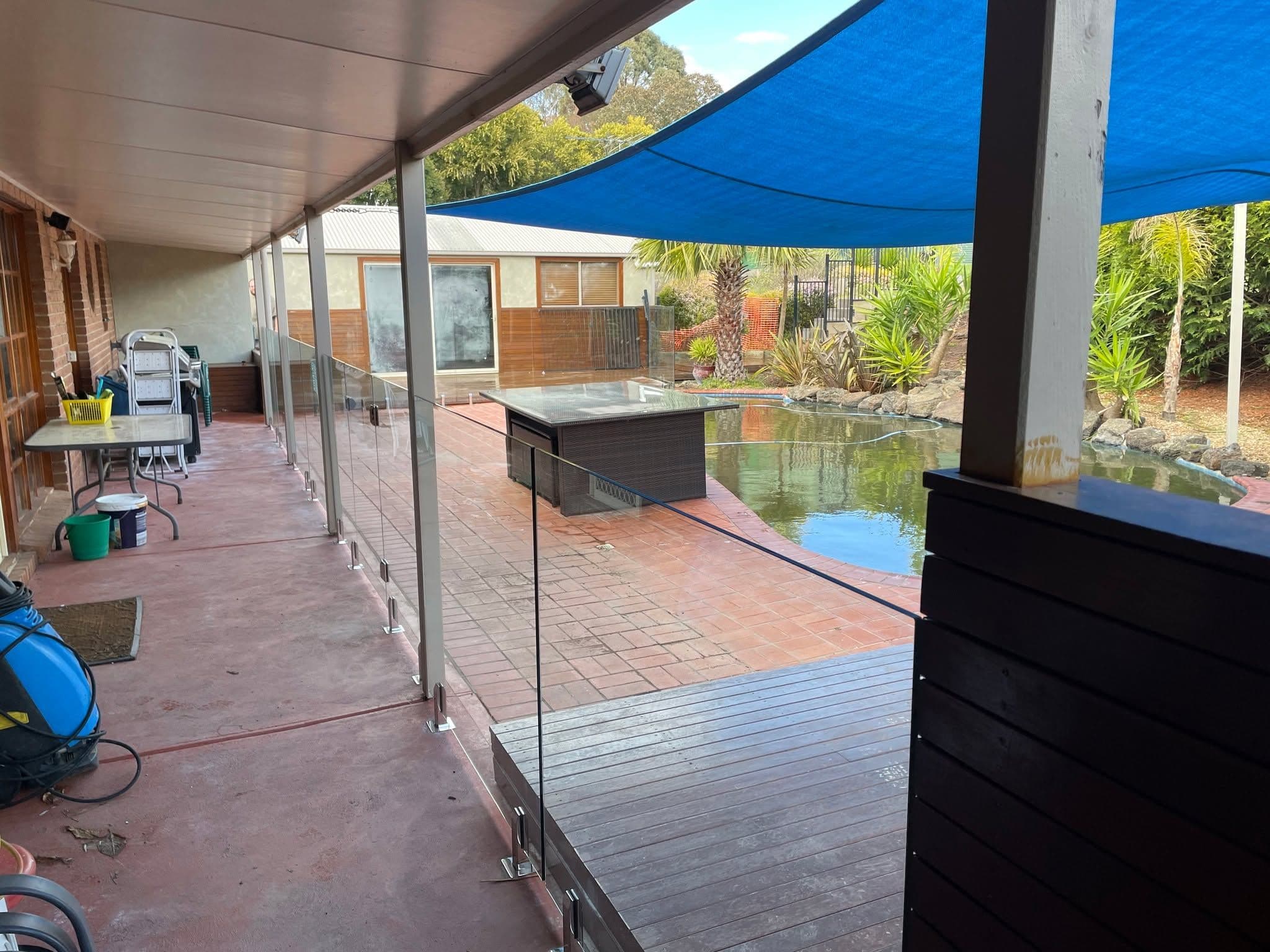 Frameless glass pool fence with chrome spigots installed along a brick patio and timber deck edge, surrounding a backyard pond/pool area with visible water.