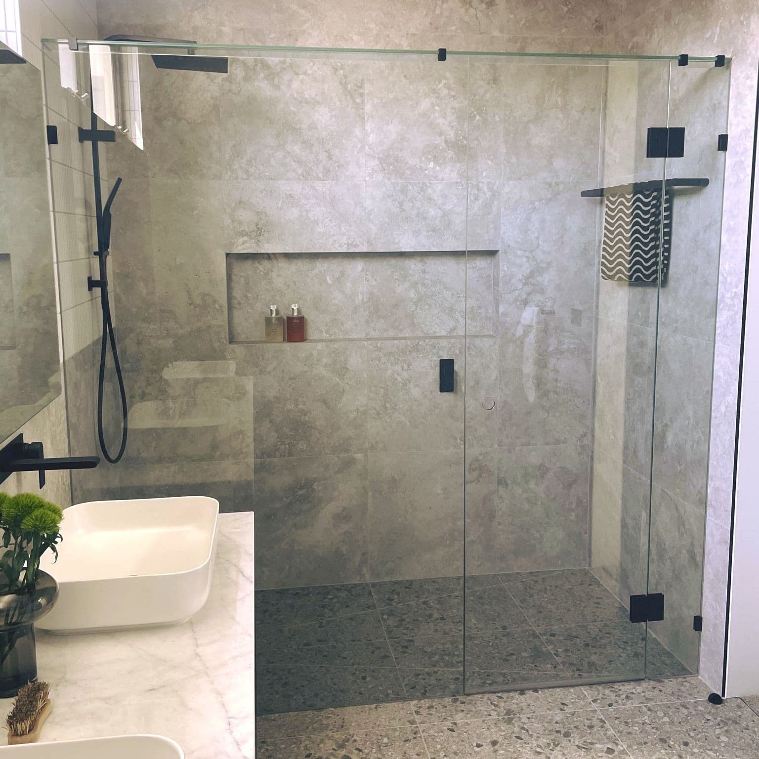 Frameless shower enclosure in a 90-degree corner configuration with matte black hinges, clips, and square wall brackets on clear glass, set in a stone-tiled ensuite with a vessel sink vanity.