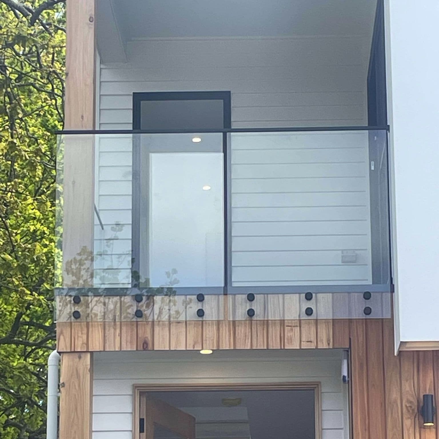 Clear glass balustrade with matte black top rail and standoff button fixings mounted on a timber-clad first-floor balcony of a modern two-storey home exterior.
