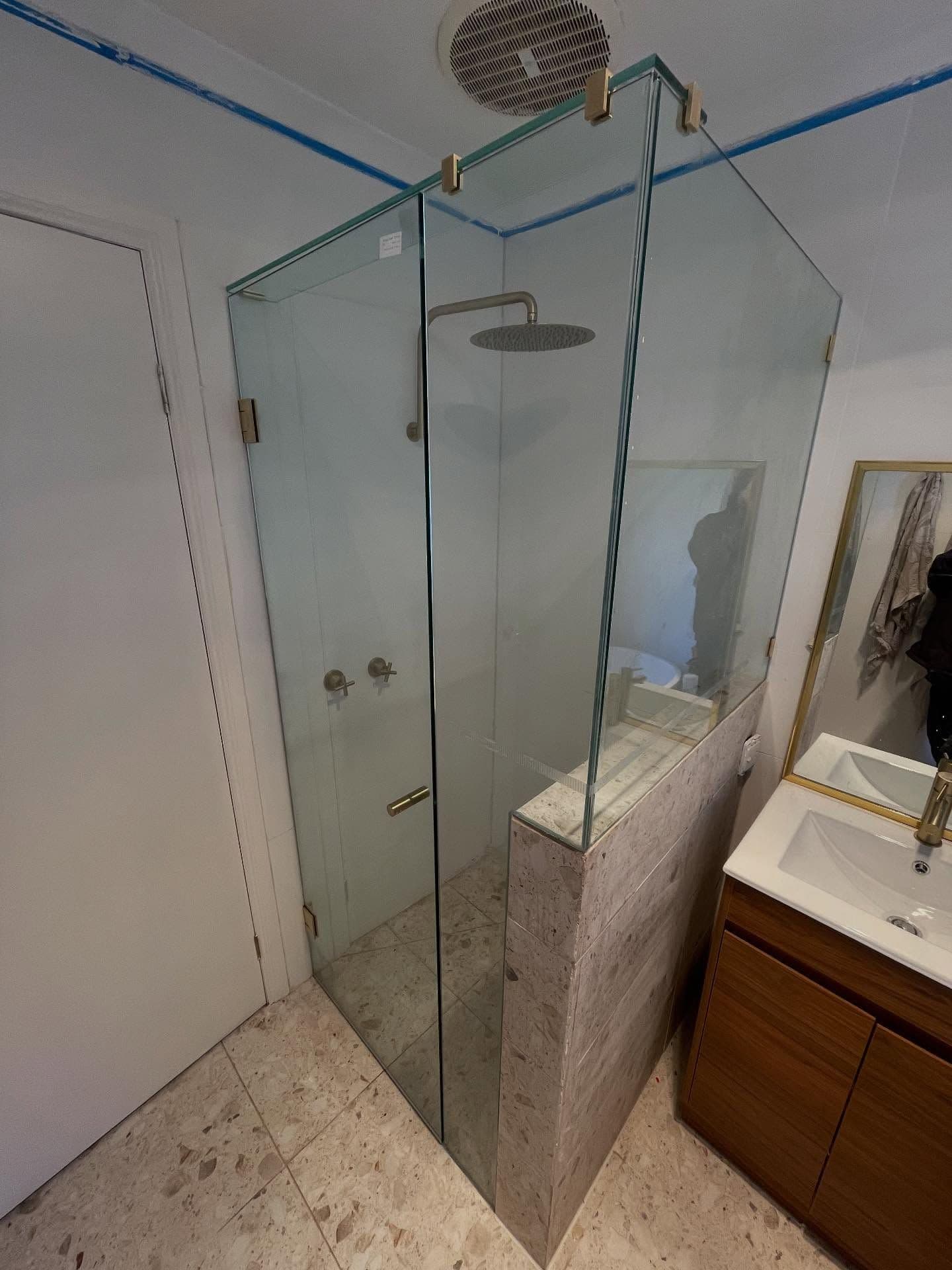 Frameless shower enclosure in a corner configuration with brushed gold hinges and door handle, clear glass panels mounted directly on glass edges, in a modern bathroom with terrazzo-style tiling and timber vanity.