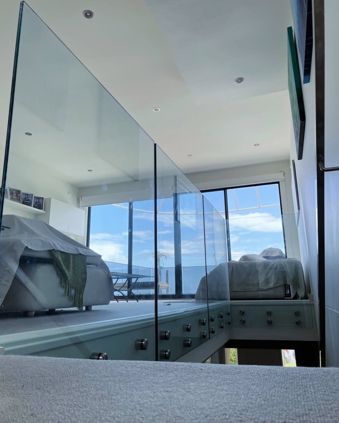 Frameless glass balustrade with chrome spigots mounted in a channel along a mezzanine or upper-floor staircase opening, overlooking an interior living space with floor-to-ceiling windows and ocean views beyond.