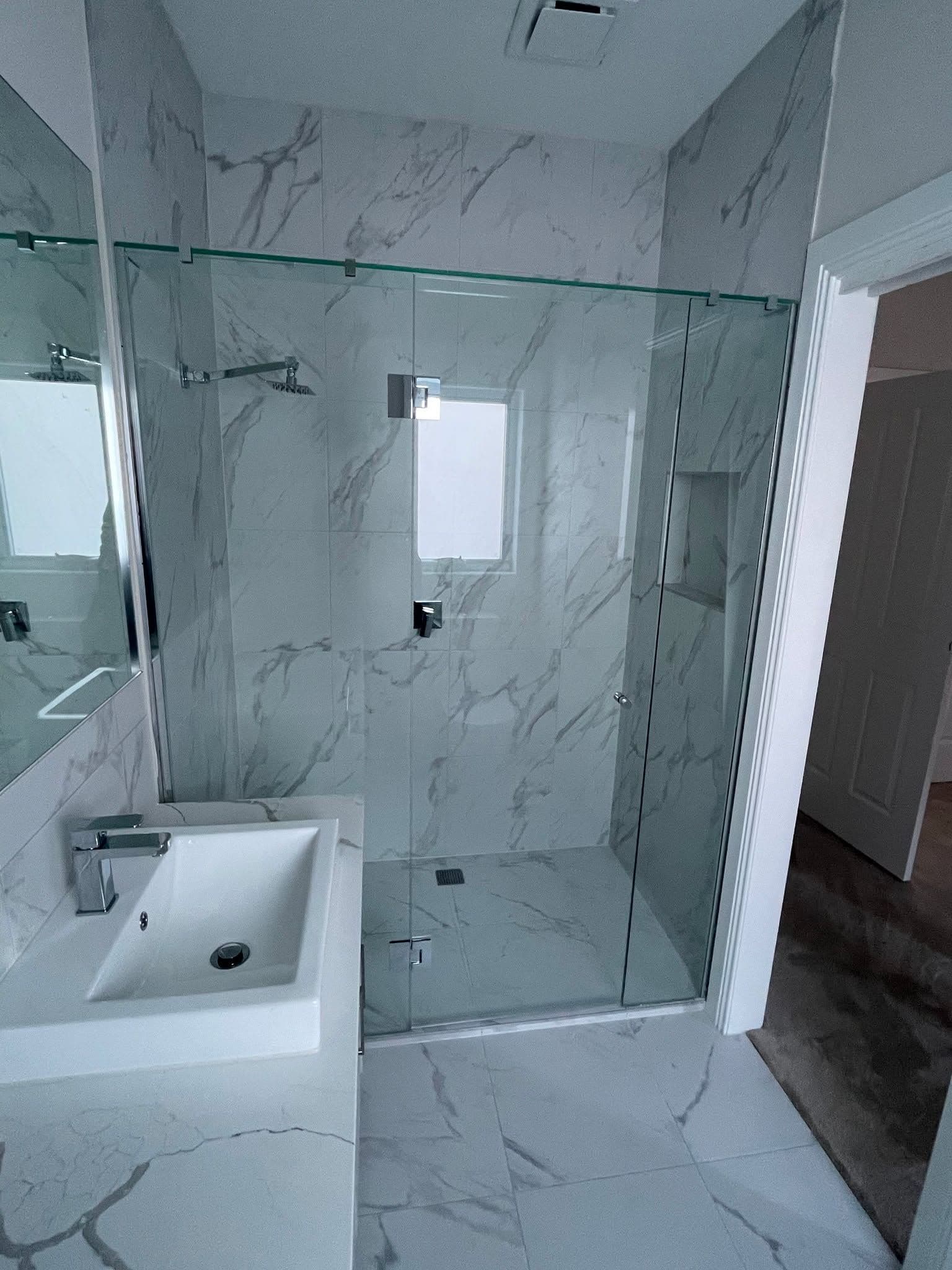 Semi-frameless shower enclosure with a sliding door on a visible top aluminium rail and chrome hardware, set in a luxurious marble-look tiled bathroom with a white basin vanity.