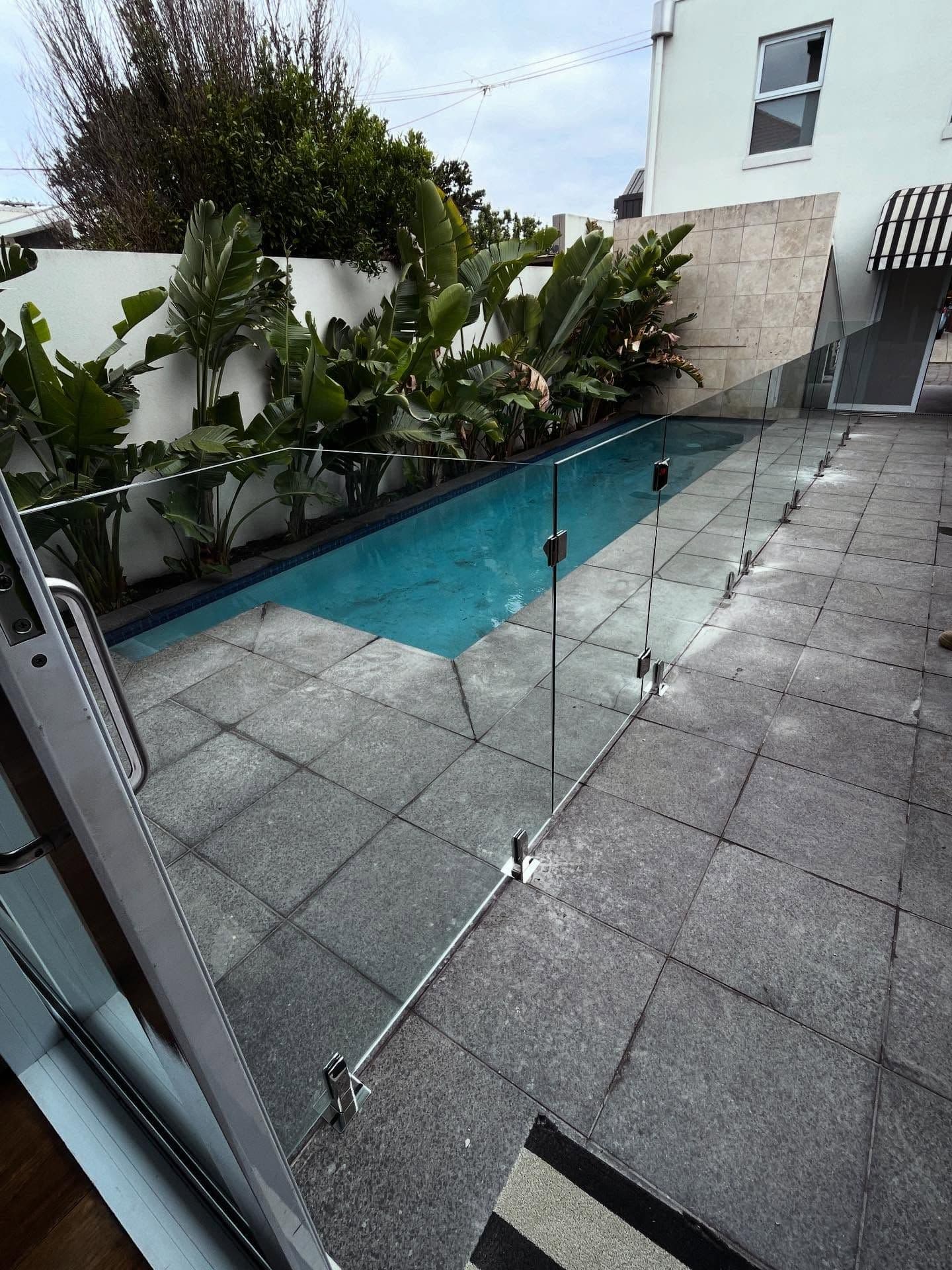 Frameless glass pool fence with chrome spigots core-drilled into grey stone coping surrounding a narrow lap pool in a tropical courtyard setting.