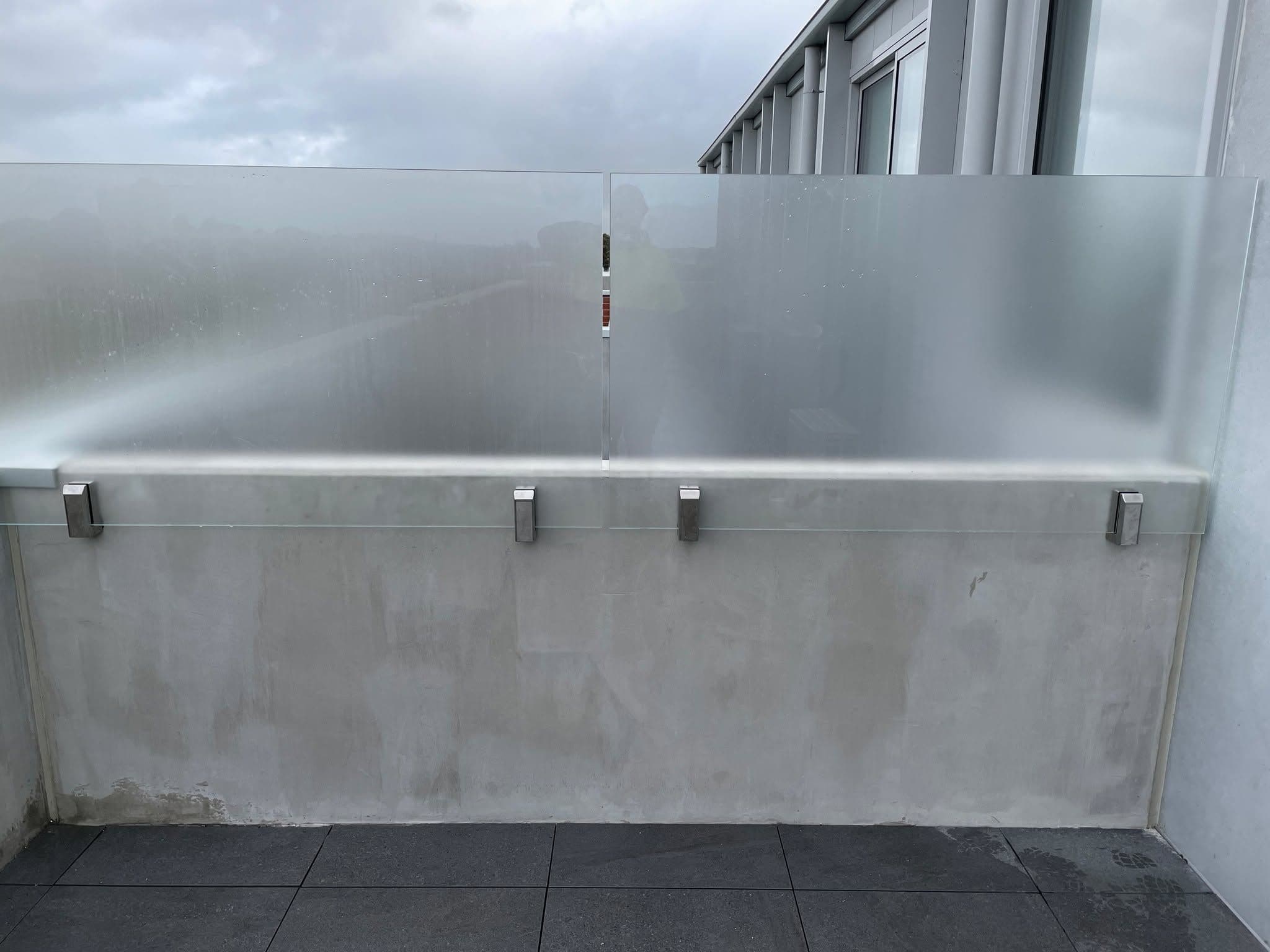 Acid etched glass balustrade panels mounted with brushed nickel standoff clamps to a rendered parapet wall on an elevated rooftop terrace or balcony.