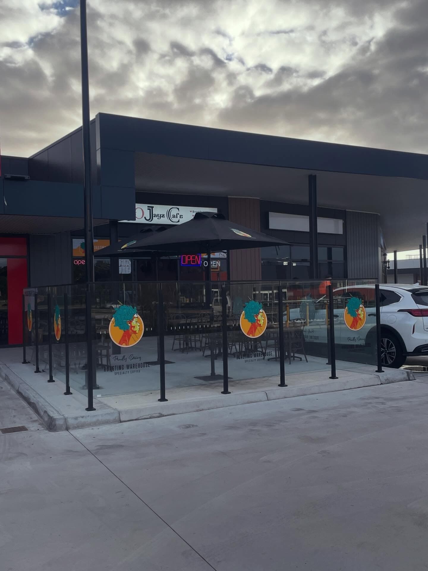 Clear glass balustrade panels with matte black spigot posts forming an outdoor alfresco dining enclosure at a commercial café, with branded vinyl graphics applied to the glass.