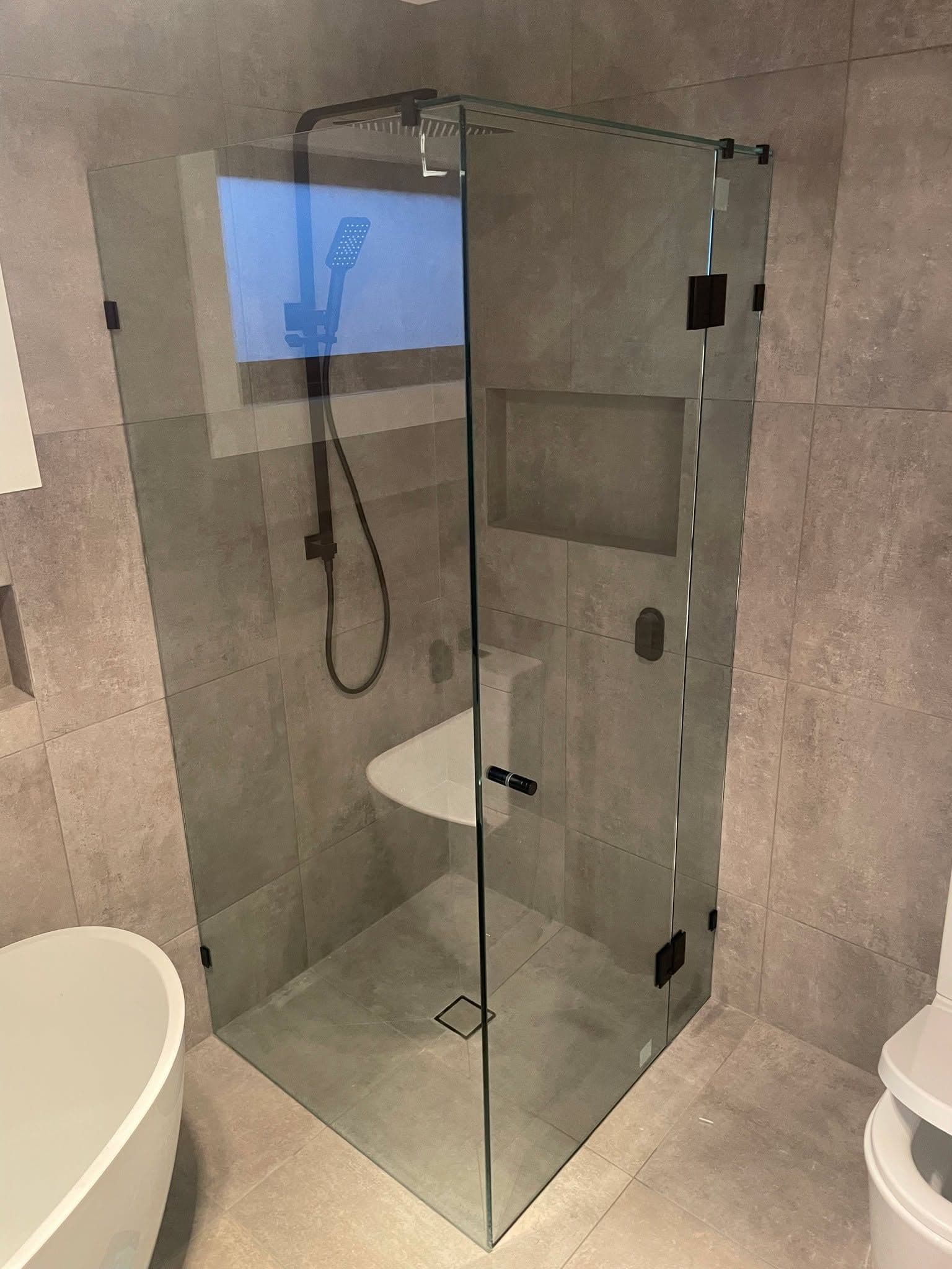 Frameless shower enclosure in a corner configuration with matte black square hinges and clips, clear glass panels, and a hinged door in a concrete-look tiled bathroom.