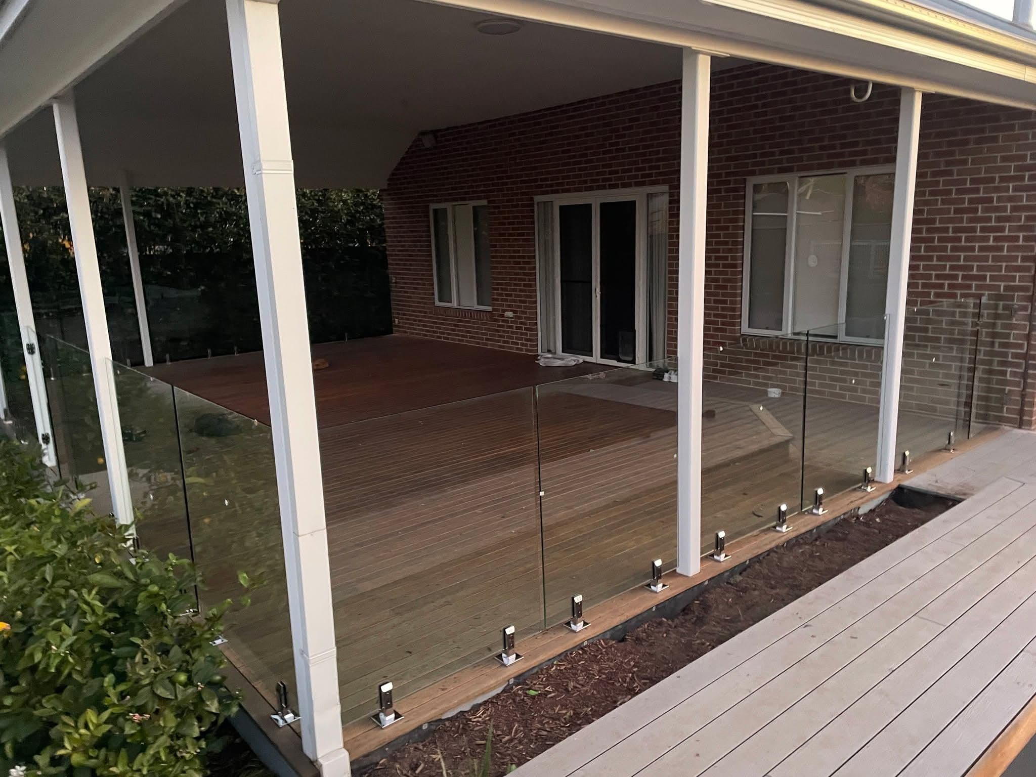 Frameless glass balustrade with chrome square spigots core-drilled into timber decking along the perimeter of a covered alfresco entertaining area adjacent to a brick home.
