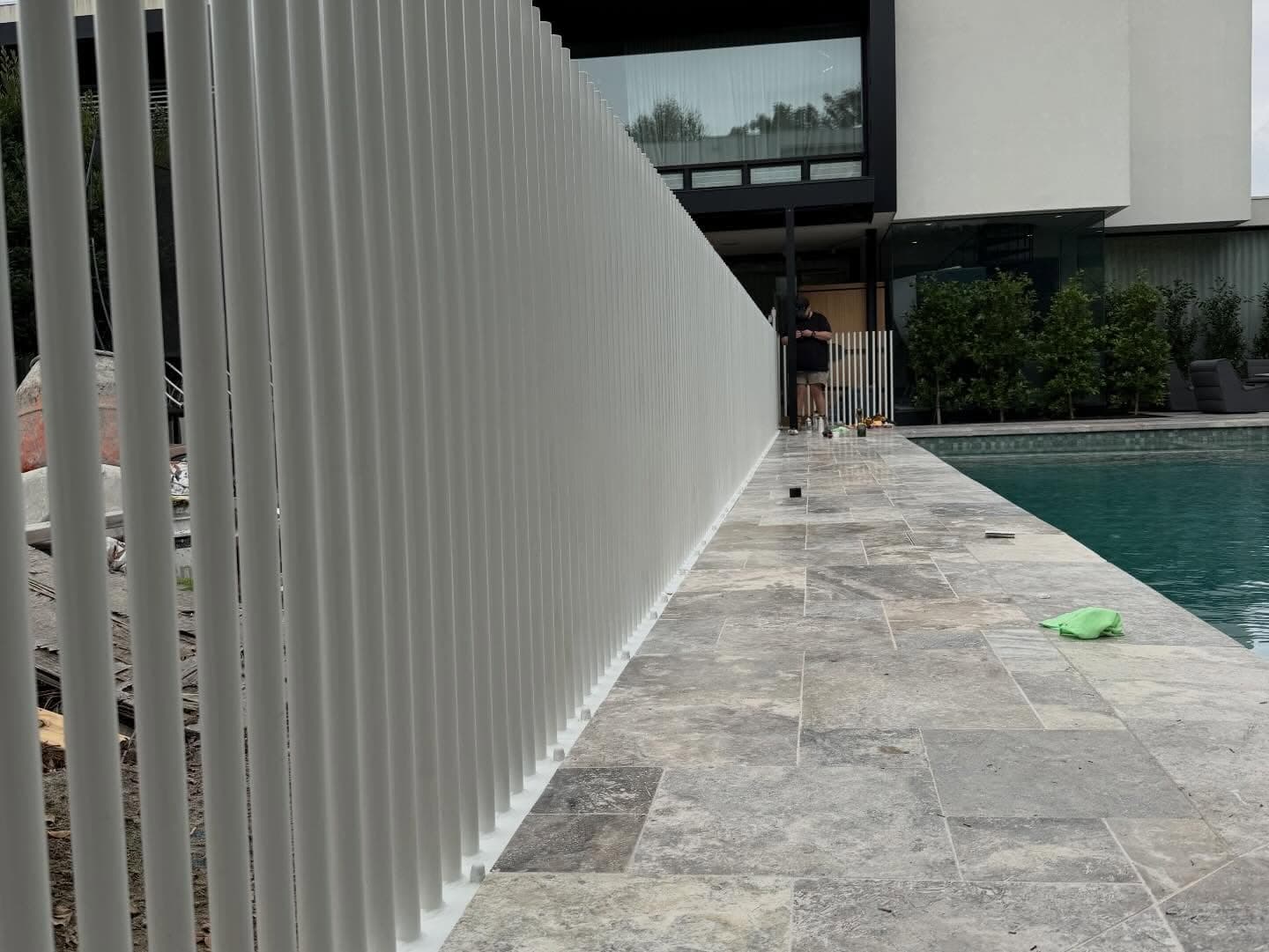 Reeded/fluted glass pool fence panels installed along a stone-paved pool coping beside a modern residential swimming pool, with white spigot bases visible at the bottom of each panel.