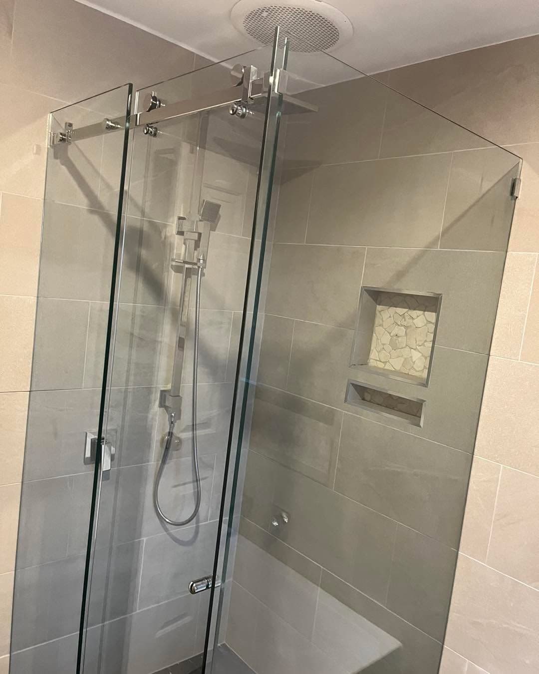 Frameless shower enclosure in a corner configuration with chrome hinges and clips mounted directly onto clear glass, set in a neutral large-format tiled bathroom with a mosaic niche feature.