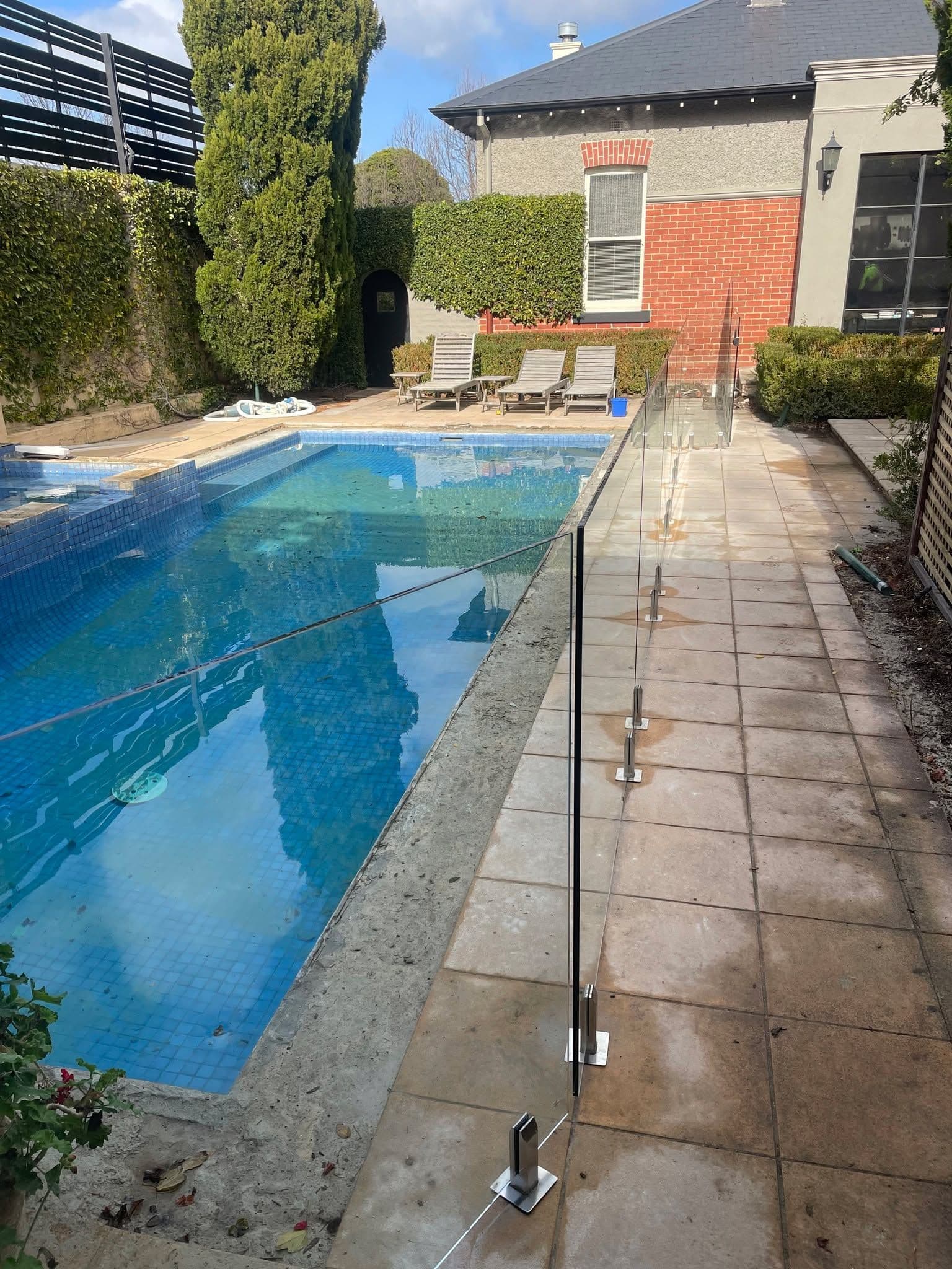Frameless glass pool fence with chrome spigots core-drilled into concrete coping alongside a tiled residential swimming pool, with a classic brick home and landscaped garden in the background.