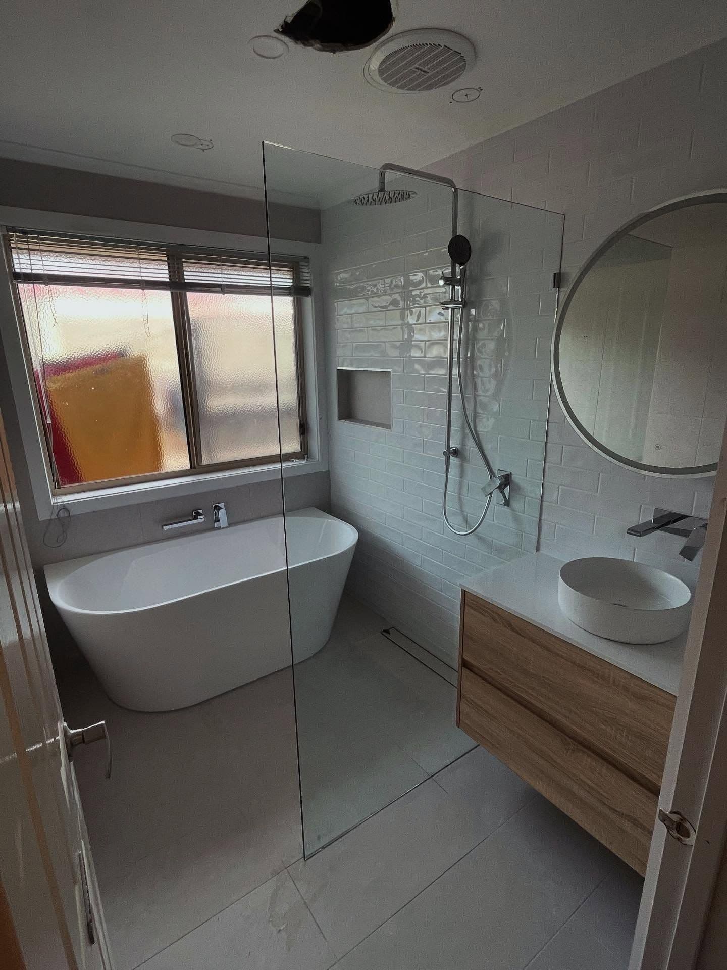 Frameless shower screen fixed as a single clear glass panel with no visible frame or hinges, forming an open wet area beside a freestanding bath in a modern white-tiled bathroom with timber vanity.