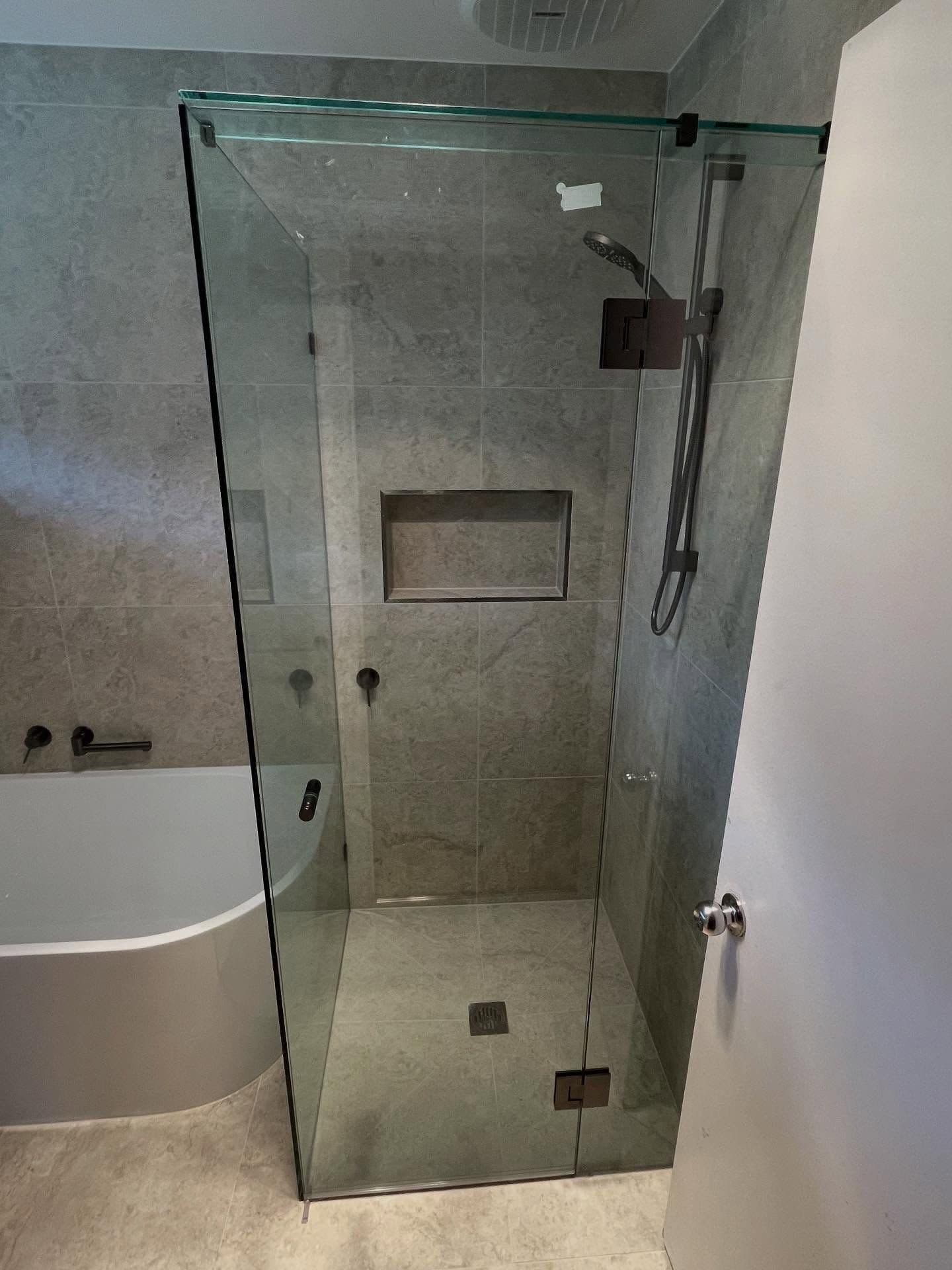Semi-frameless shower enclosure with a visible top rail/sliding track in matte black finish, corner configuration with clear glass panels, set in a grey stone-tiled bathroom alongside a freestanding bath.