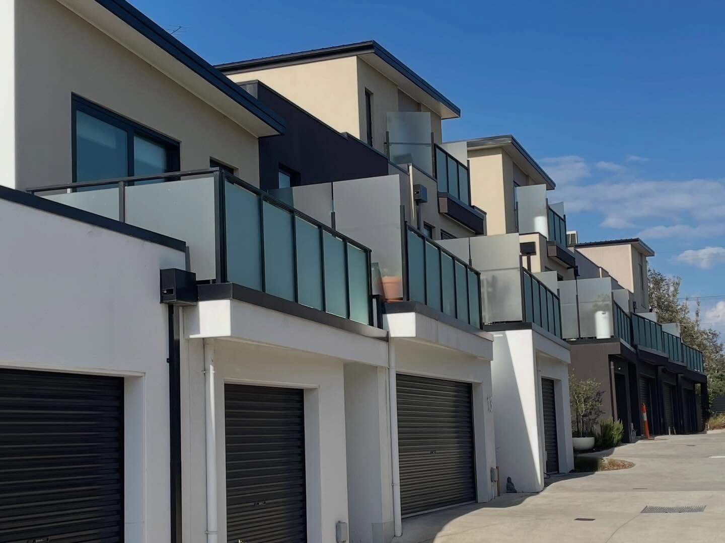 Semi-frameless glass balustrade panels with dark aluminium top rail and framing channels on first-floor balconies of a modern multi-unit residential development.