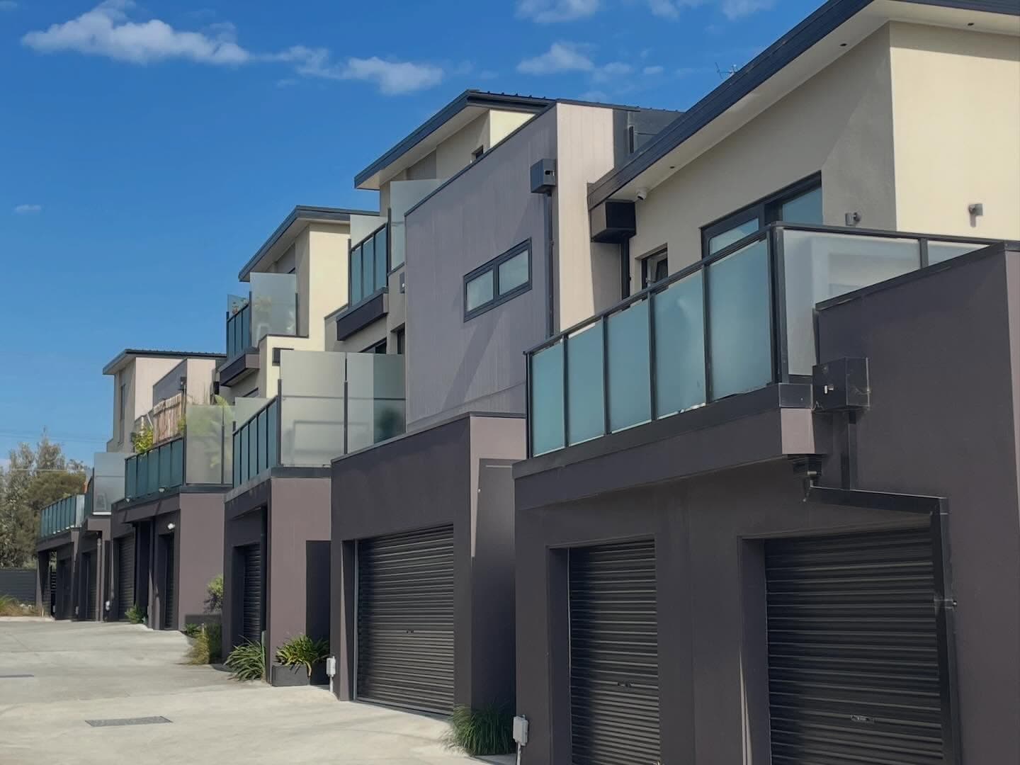 Semi-frameless glass balustrades with dark aluminium top rail and channel framing on first-floor balconies of a row of modern multi-unit townhouses, no pool visible.