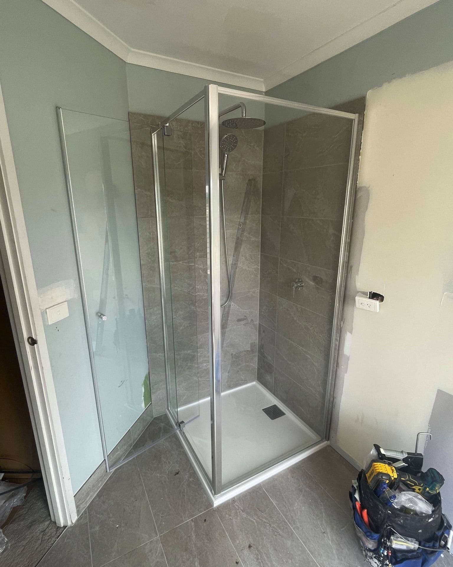 Semi-frameless shower enclosure with visible aluminium frame channels on all edges and a pivot door, chrome finish, in a grey marble-tiled bathroom under construction.