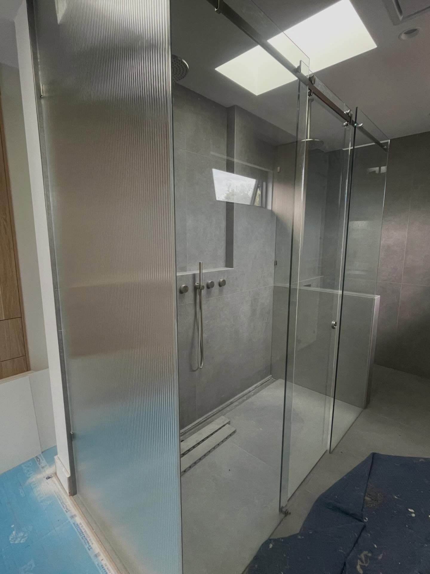 Semi-frameless shower enclosure featuring a reeded glass fixed panel and a clear glass sliding door with a visible overhead aluminium sliding track and brushed nickel hardware, set in a modern grey tiled bathroom with skylight.