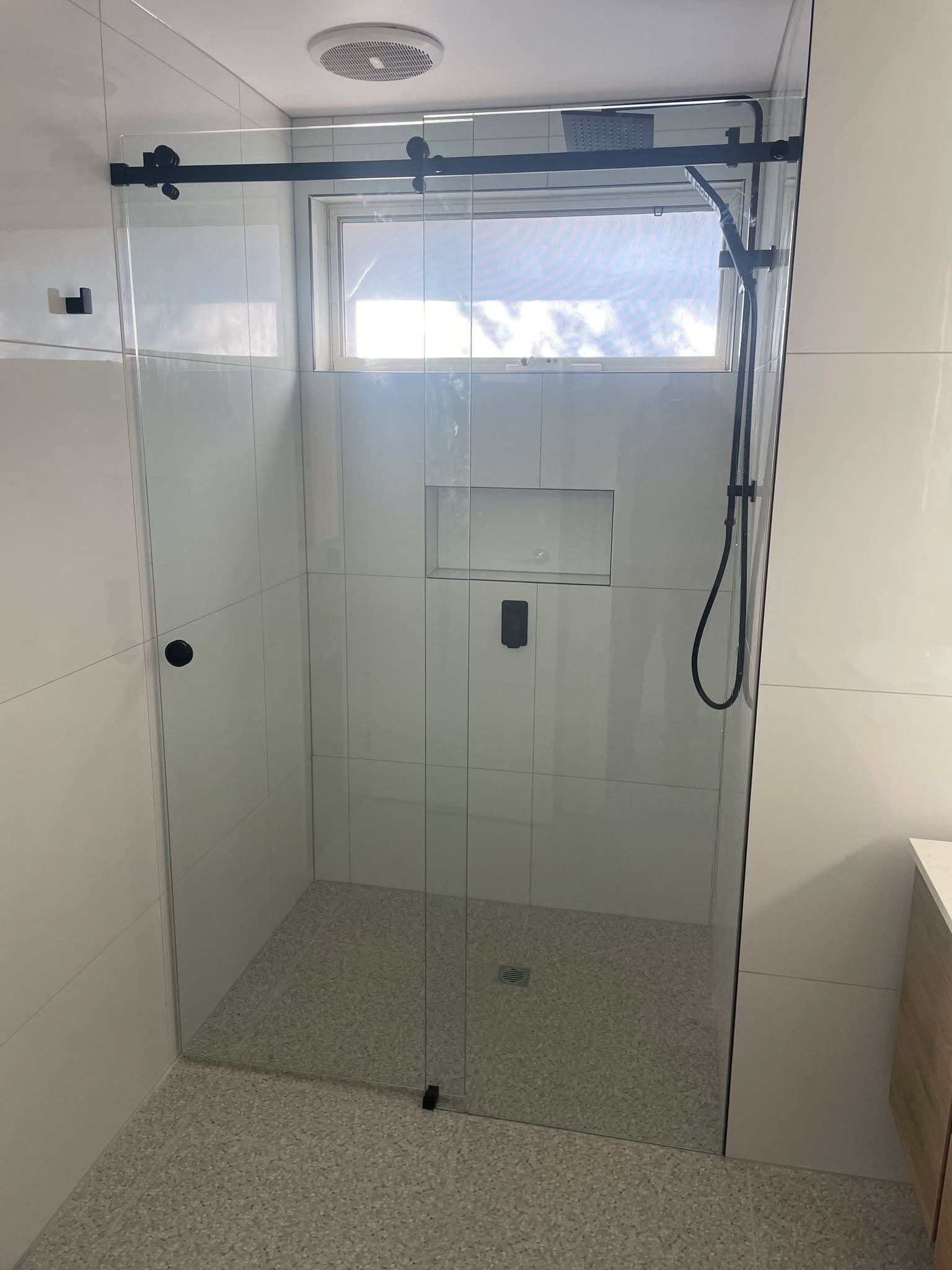 Semi-frameless sliding shower screen with a matte black top rail and sliding door track in a white tiled bathroom with terrazzo-style floor.