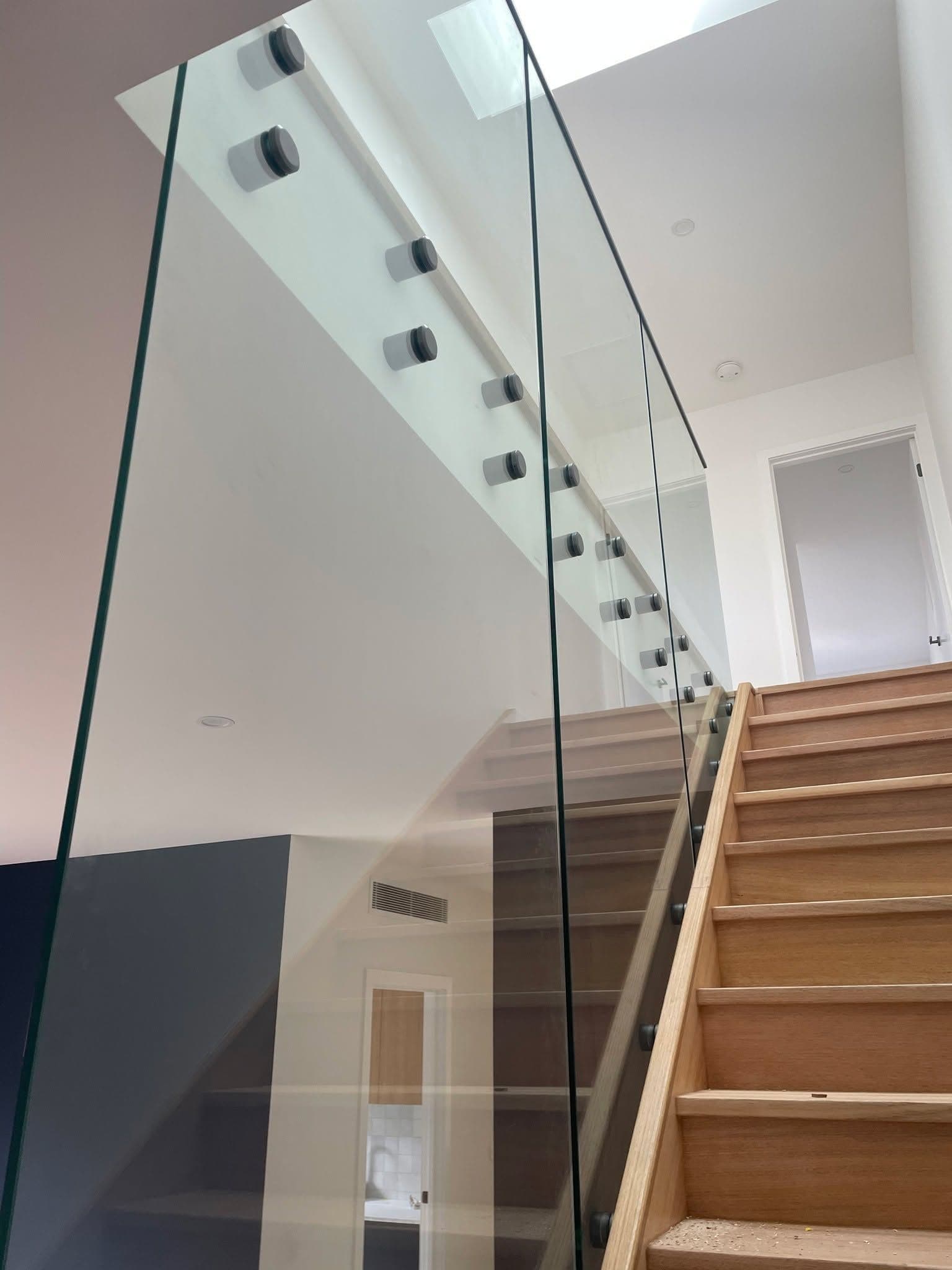 Clear glass staircase balustrade with matte black standoff fixings mounted to the wall, running alongside timber oak stairs in a modern interior home.