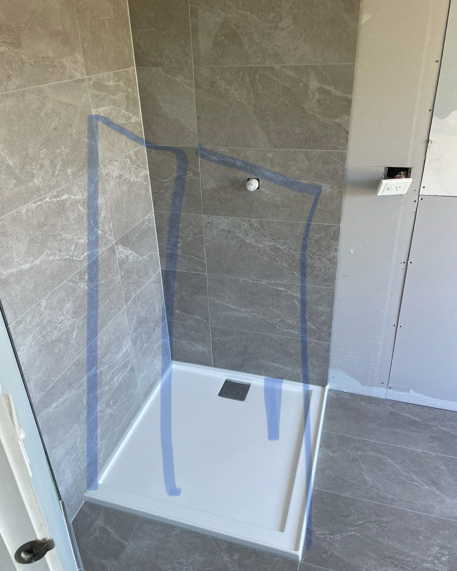 Frameless shower screen with a single clear glass panel and chrome wall hinge, installed in a partially finished bathroom with grey marble-look tiles and a white acrylic shower base.