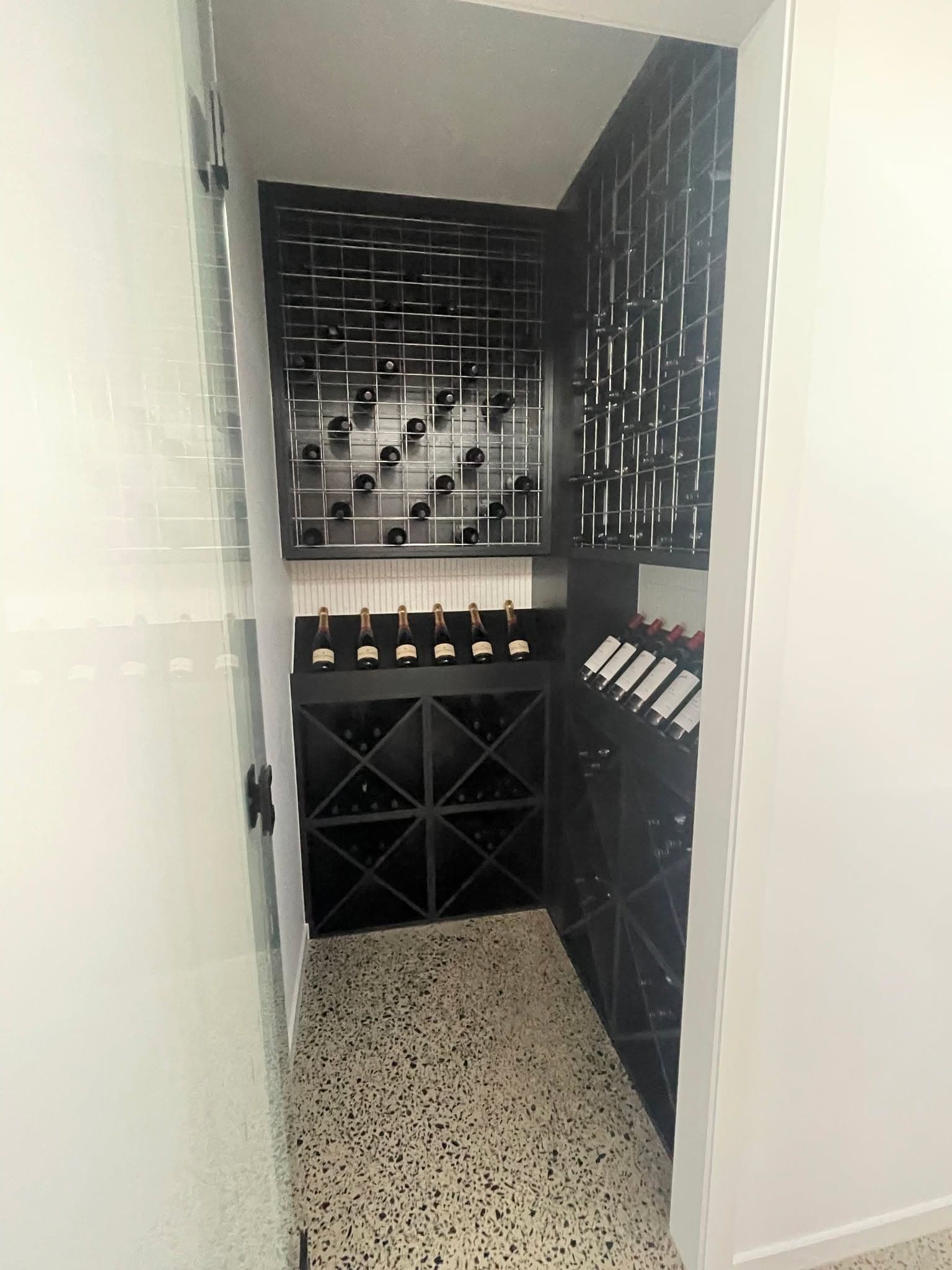 Frameless glass door with matte black hardware providing entry to a walk-in wine cellar with black wine racks and terrazzo flooring.